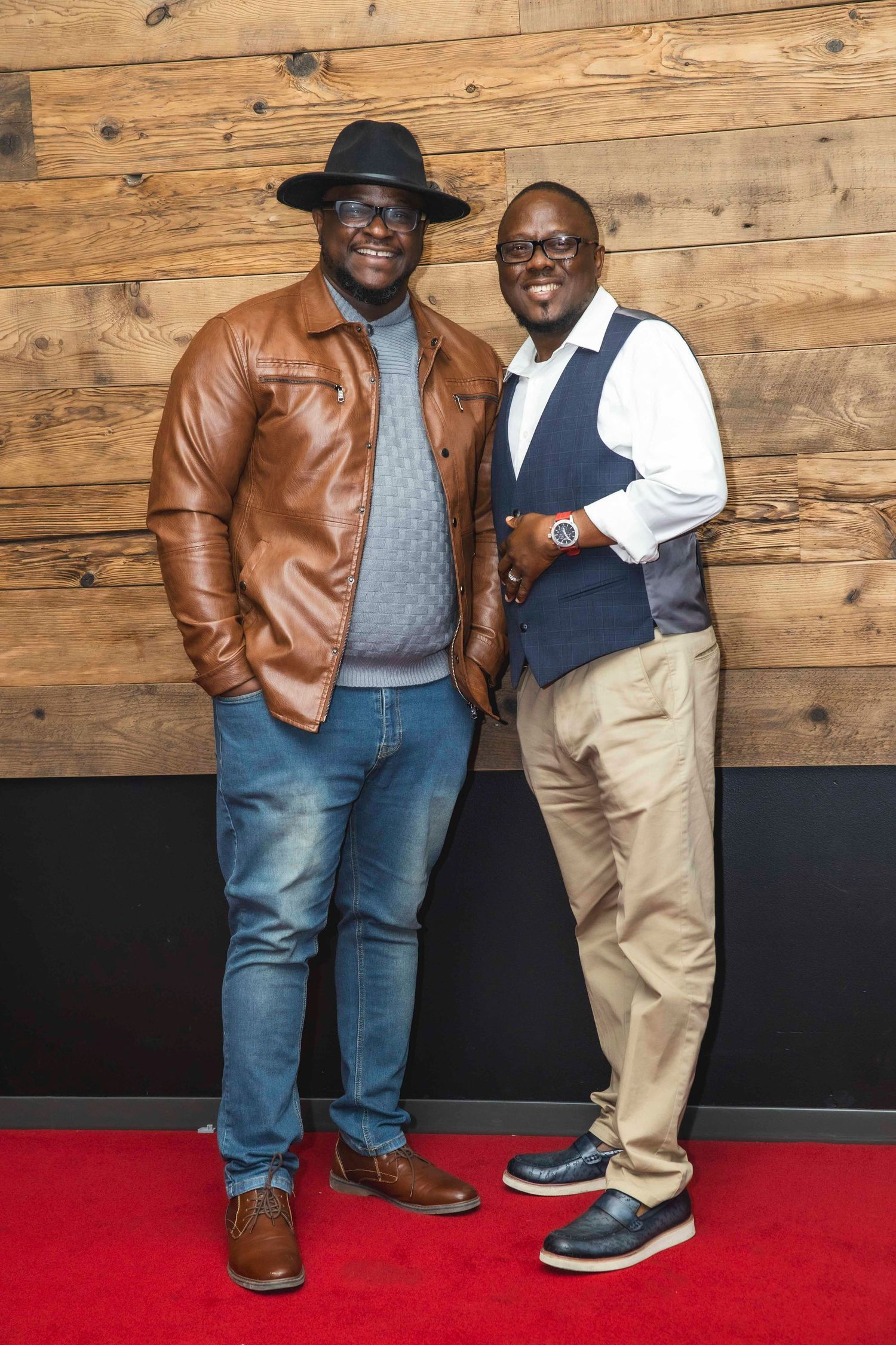Two people posing together on red carpet in front of rustic wooden wall.