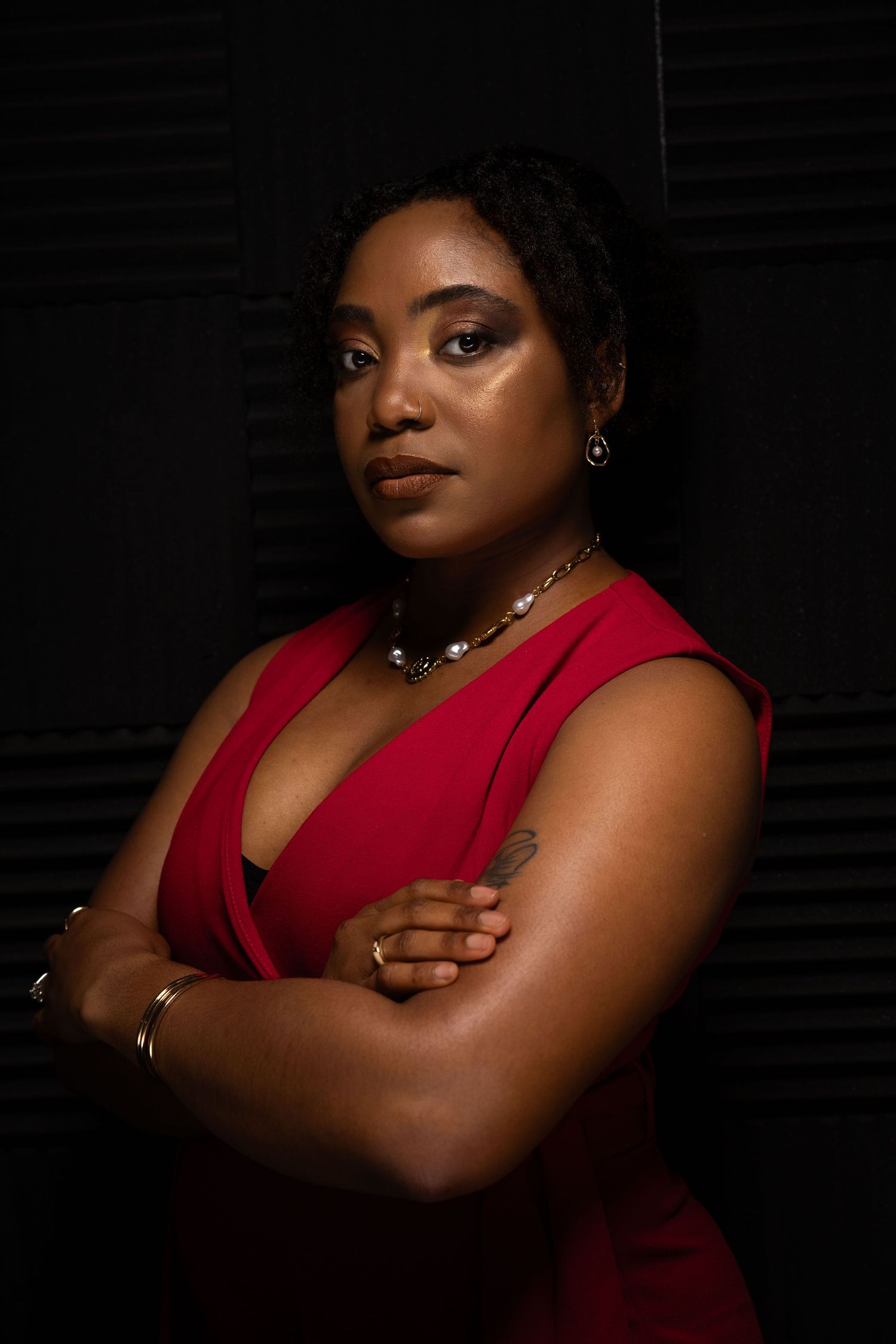 Dramatic portrait in a red sleeveless top with crossed arms against dark background.