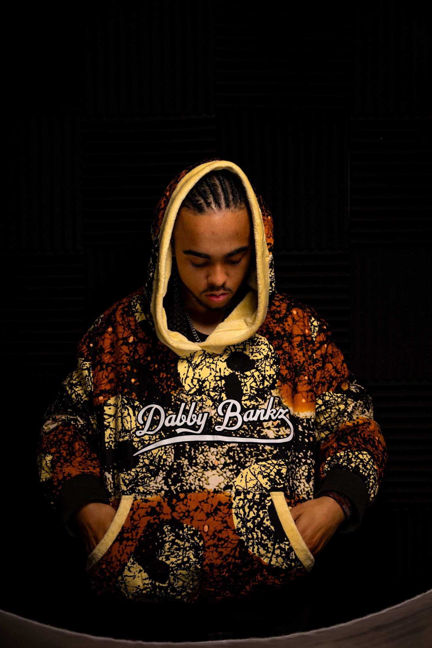 Person in patterned hoodie with braided hair against dark background.