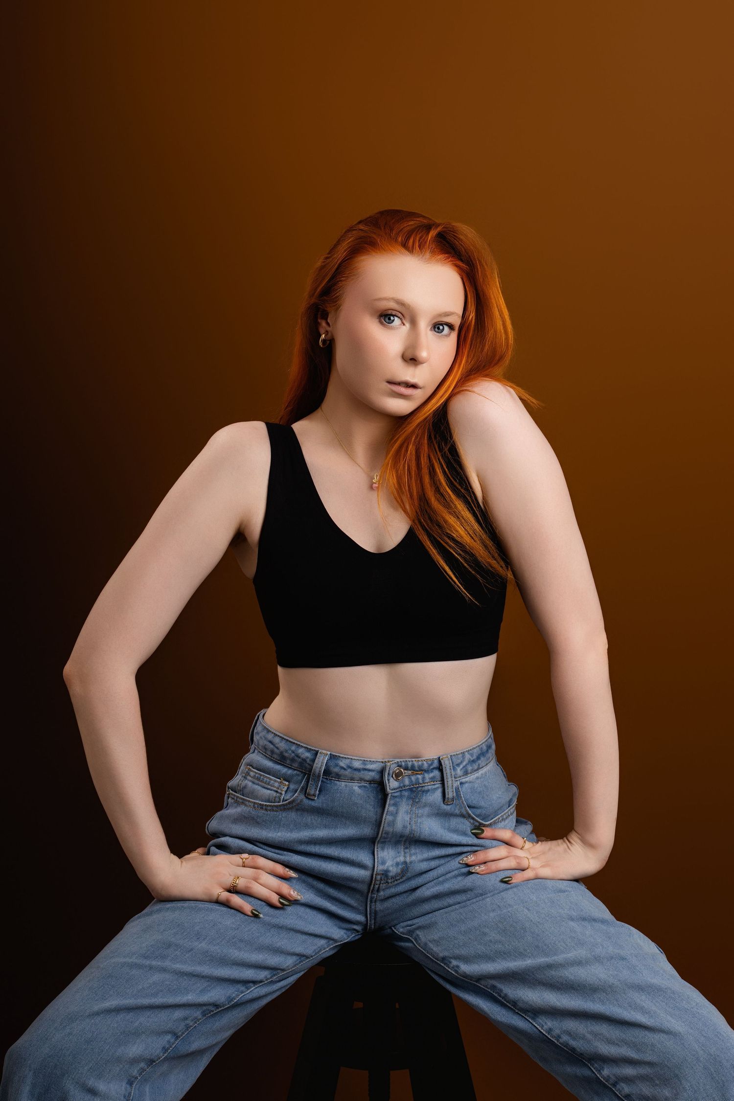 Casual fashion pose in black crop top and denim against warm brown background.
