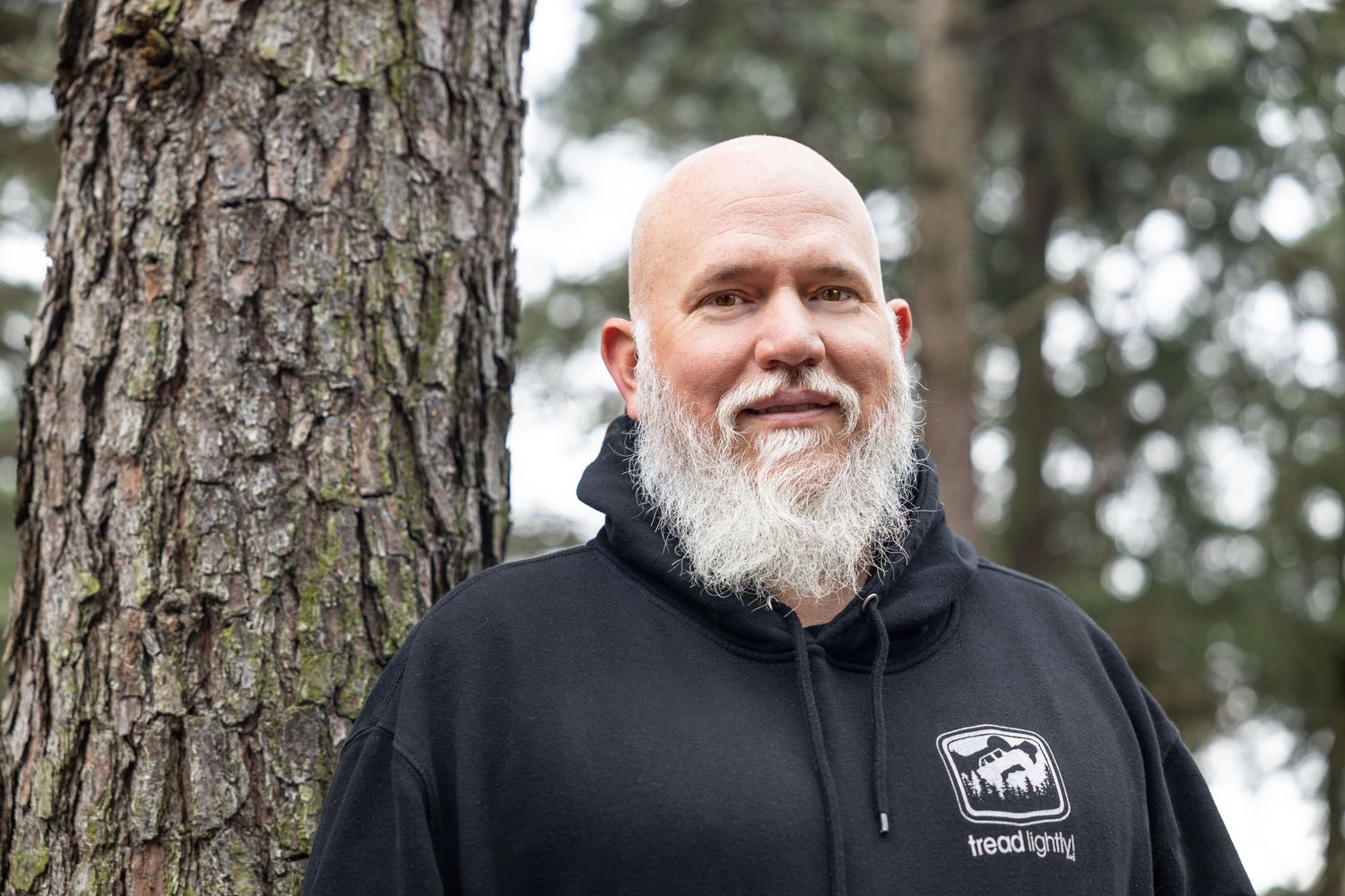 Smiling man with white beard wearing black hoodie standing next to pine tree.