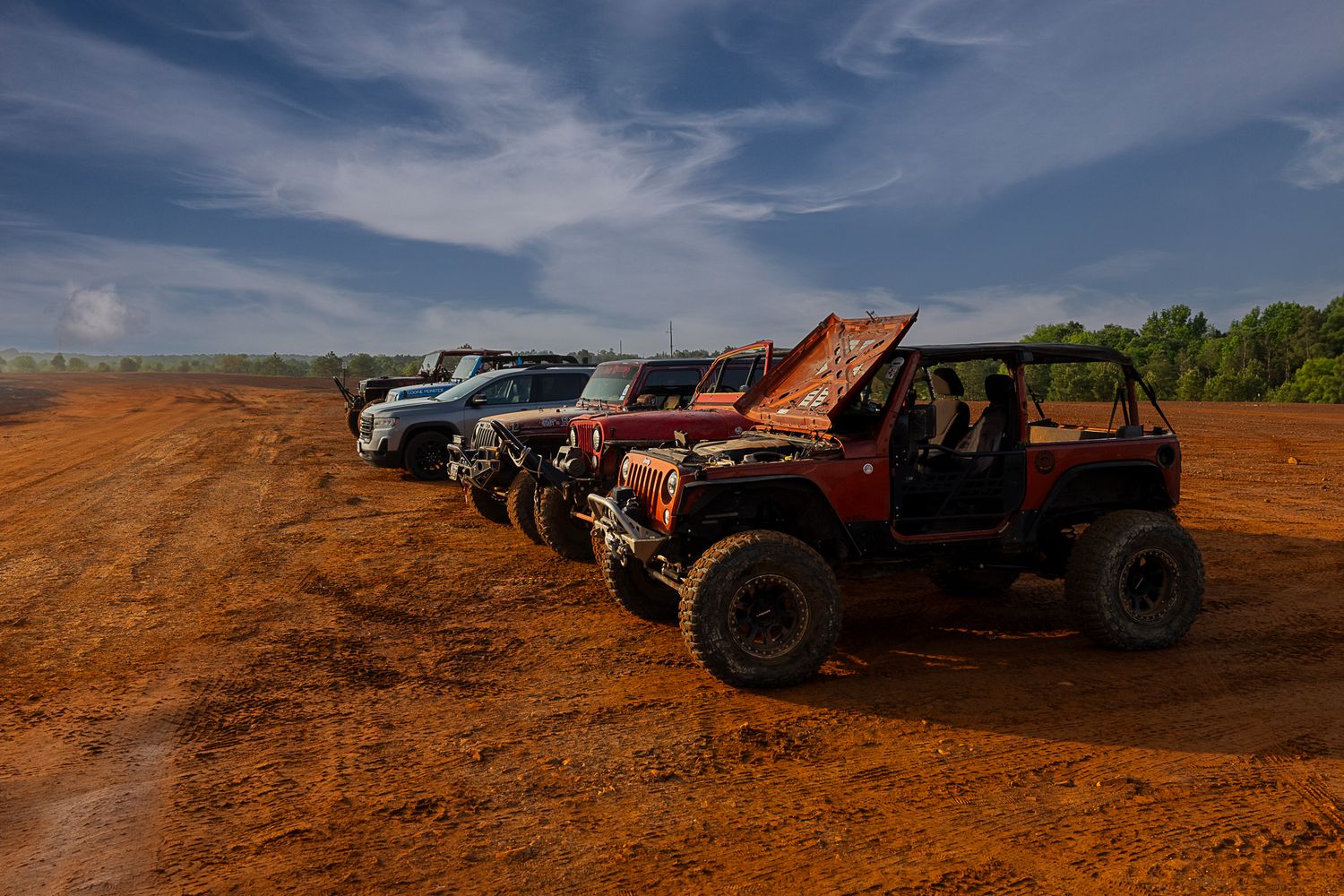 Row of modified off-road vehicles parked on red dirt under blue sky.