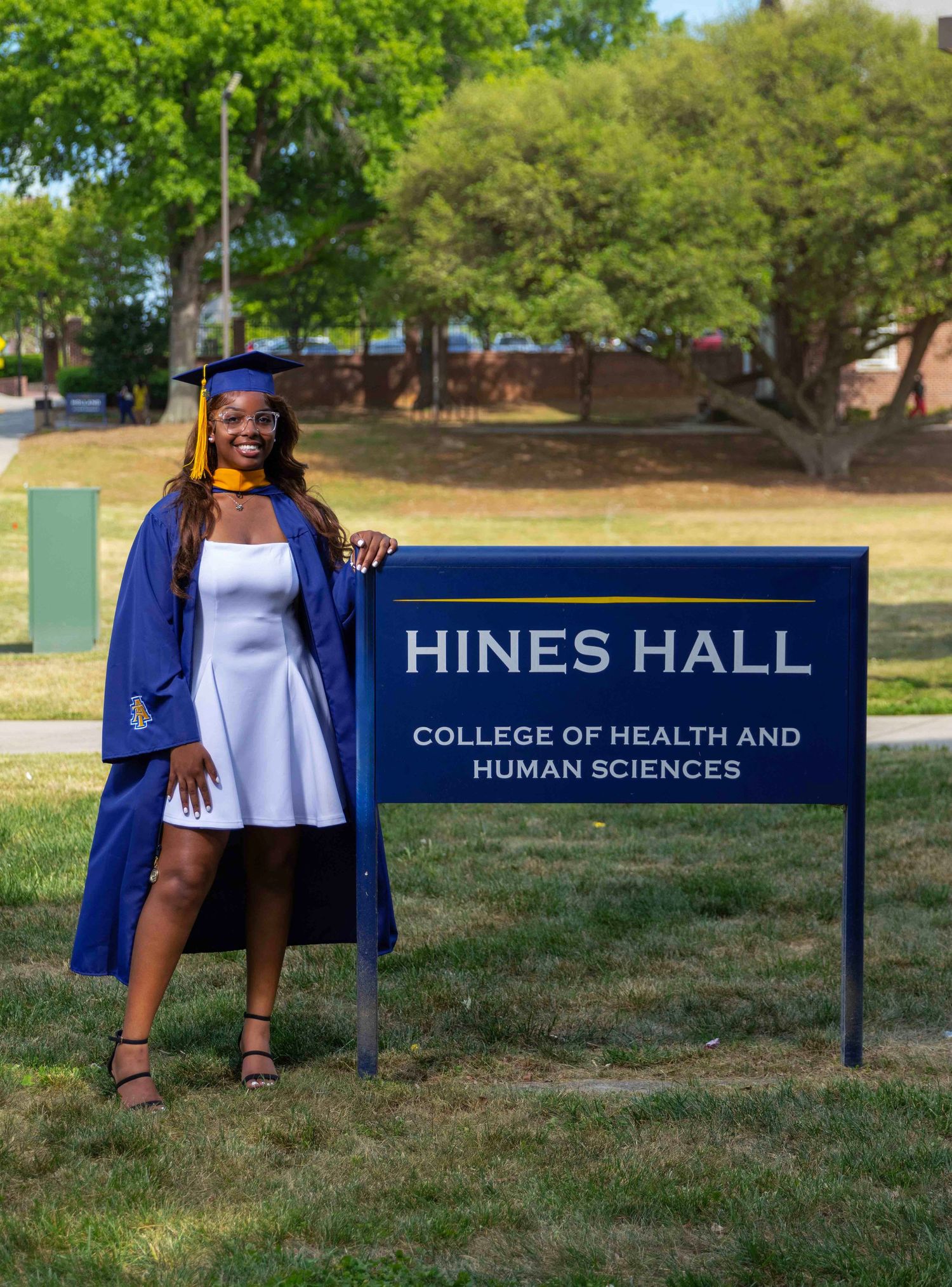 Graduate in blue cap and gown poses proudly beside Hines Hall College of Health and Human Sciences sign outdoors.