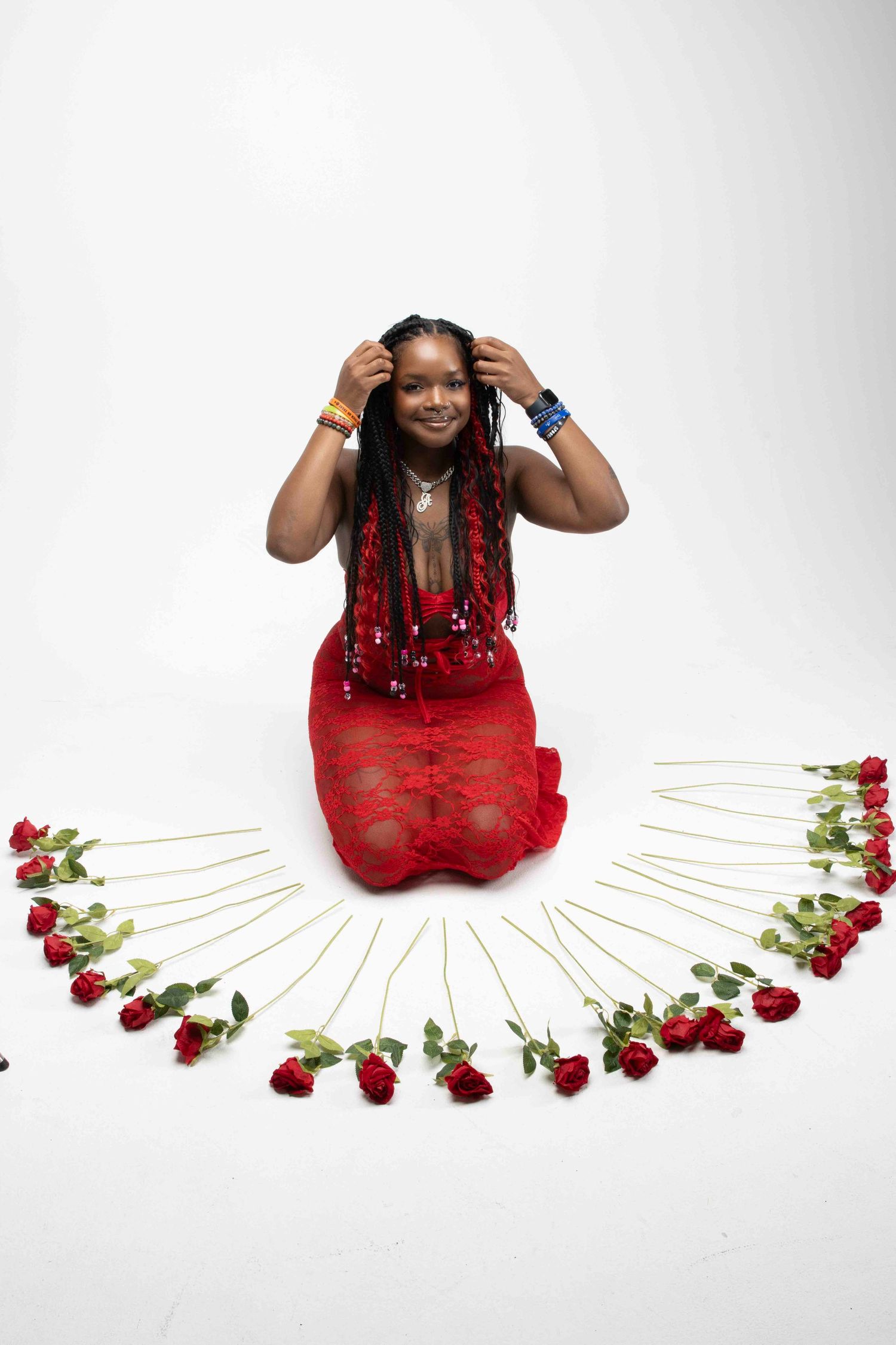 Woman in red lace dress kneeling surrounded by red roses arranged in semicircle on white background.