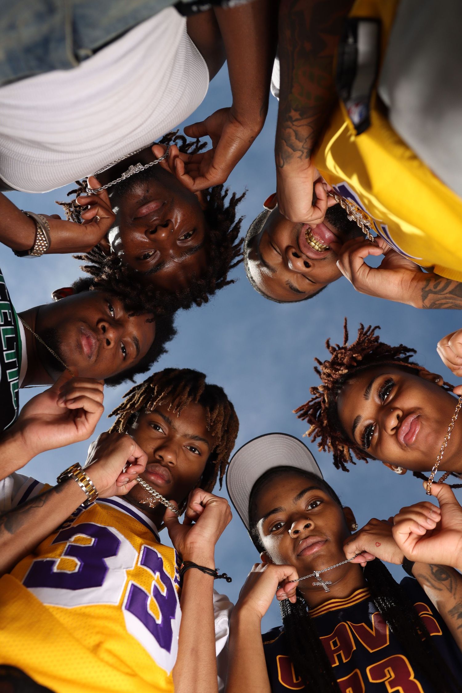 Group of young men with dreadlocks in NBA jerseys forming a circle huddle, shot from below against blue sky.