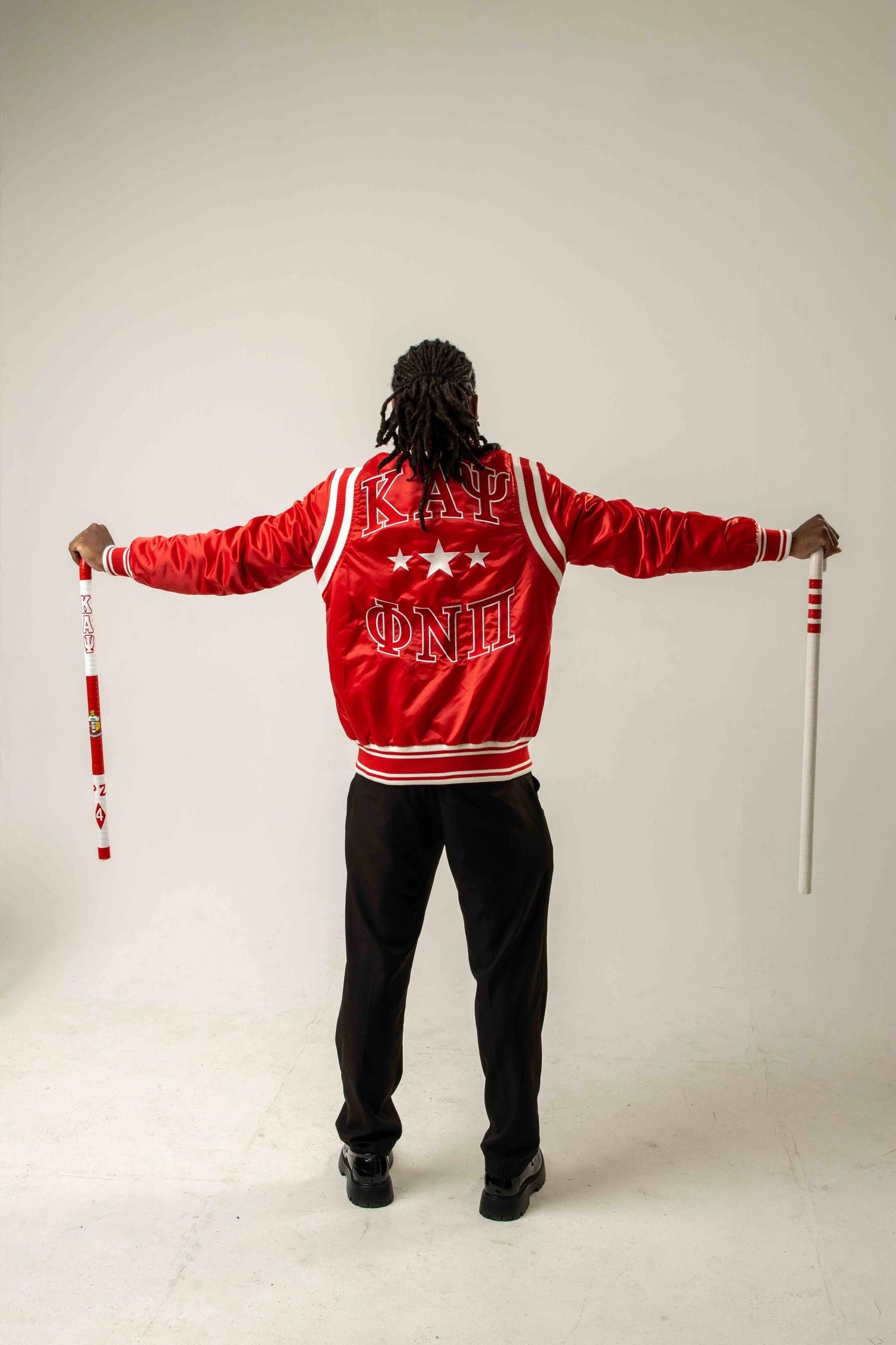 Man wearing red Kappa Alpha Psi fraternity jacket holding canes with arms outstretched, viewed from behind.