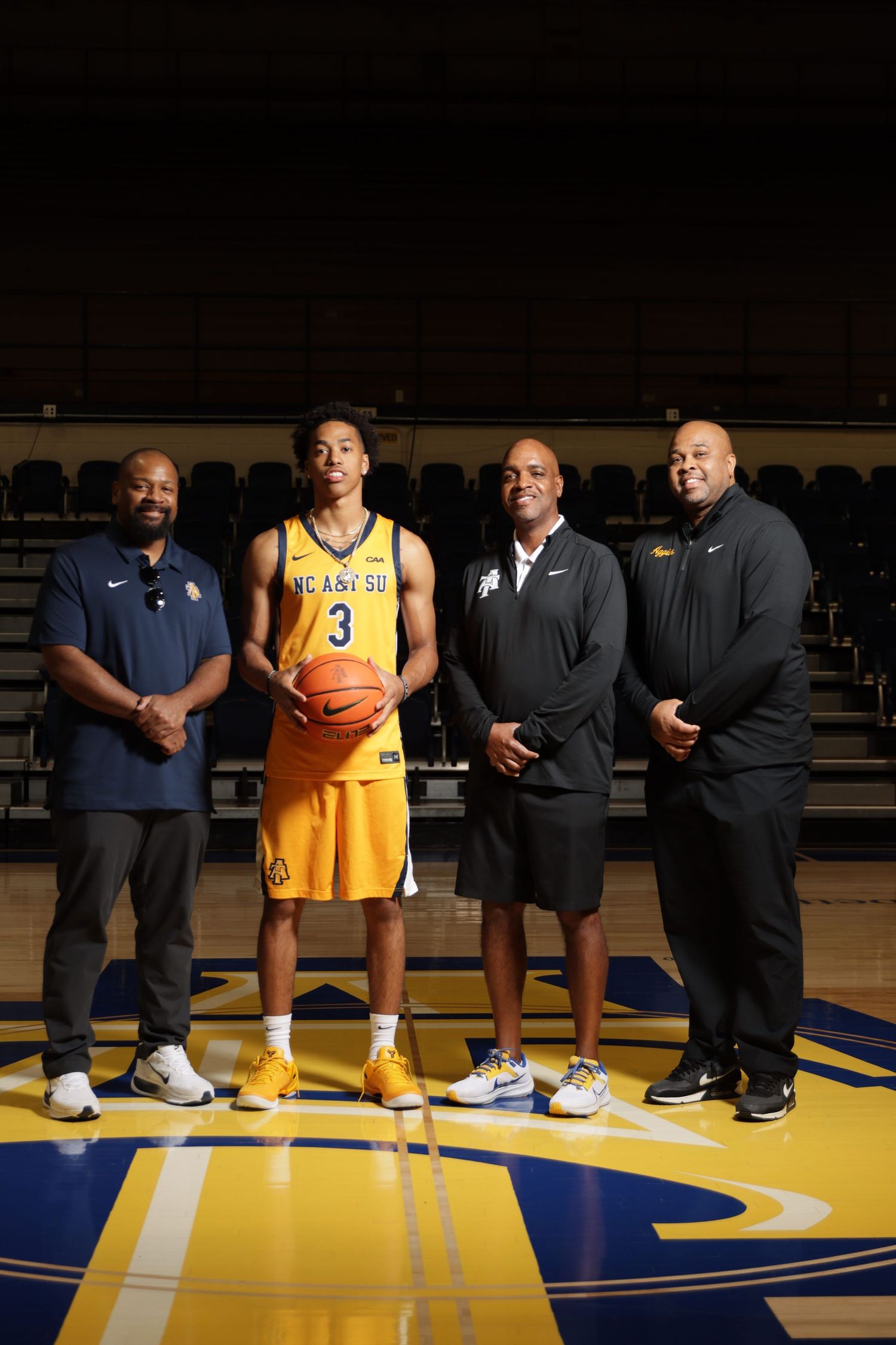 NC A&T basketball player #3 poses with three coaches on the court in yellow uniform holding a basketball.