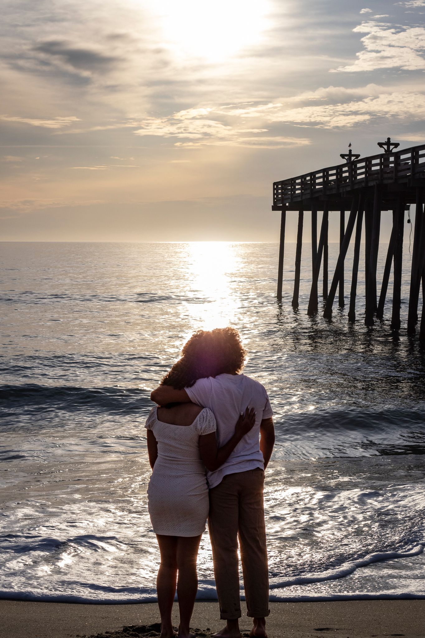 Couple embracing while watching sunset at wooden pier on beach.