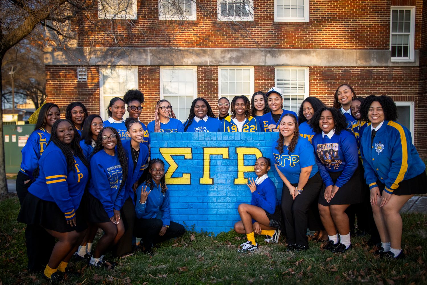 A large group wearing blue and gold poses together in front of a sorority banner on a brick building.
