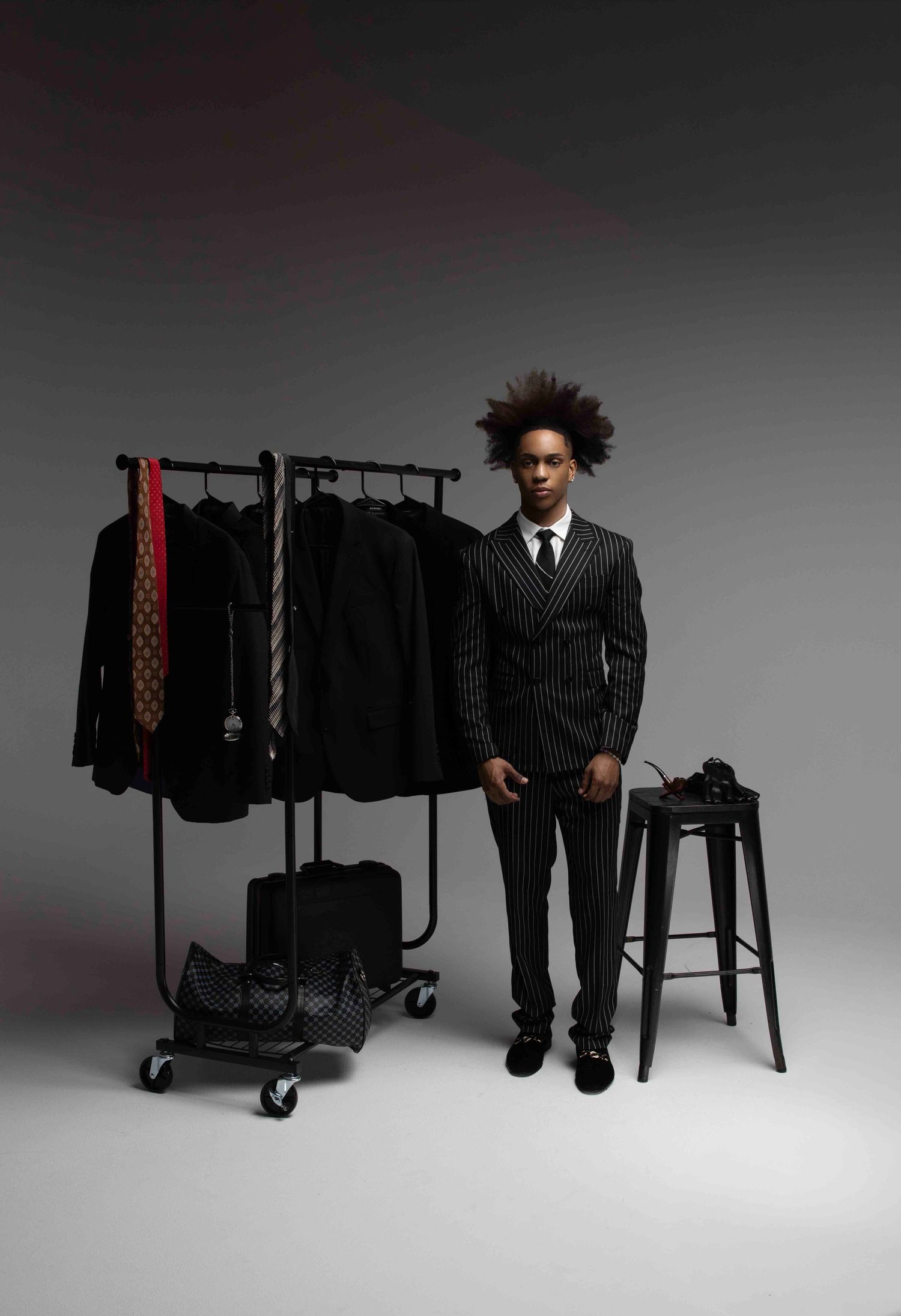 Man in pinstripe suit stands beside clothing rack and stool in studio setting.