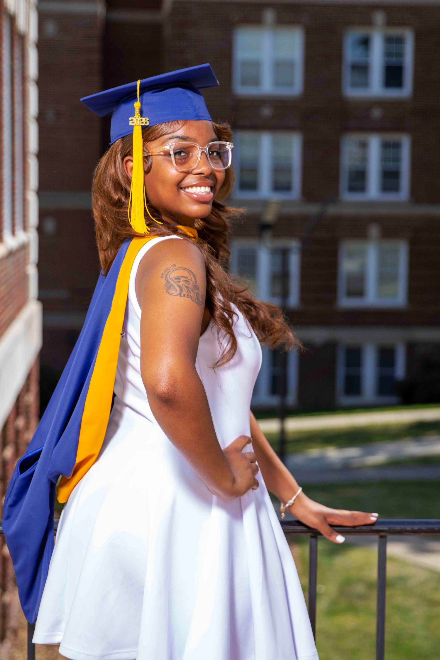 Smiling Black woman in blue graduation cap and yellow tassel wearing white dress poses on balcony near brick building.