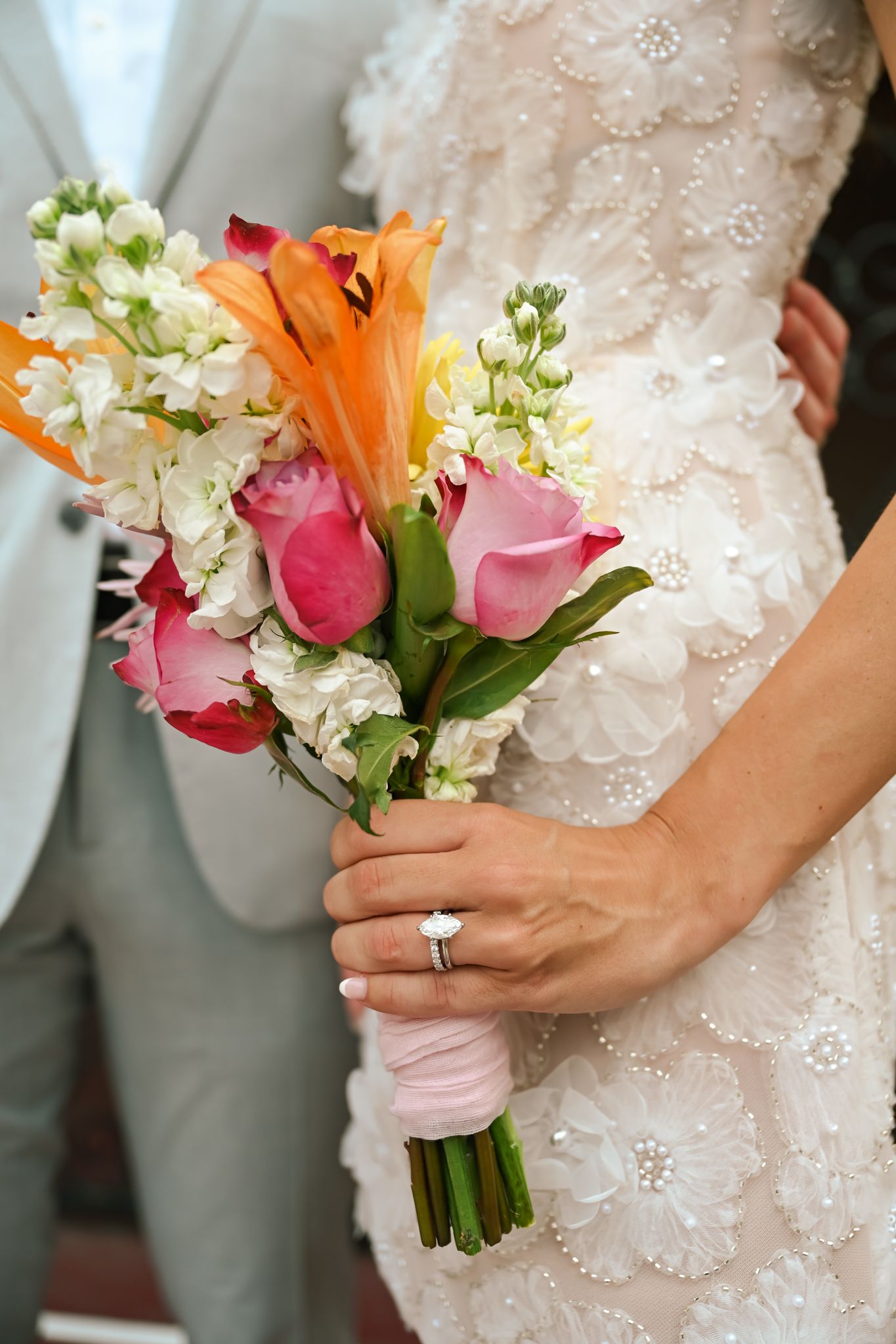 Bride in white beaded gown holds colorful wedding bouquet with pink roses, orange lilies, and white flowers, wearing diamond ring.