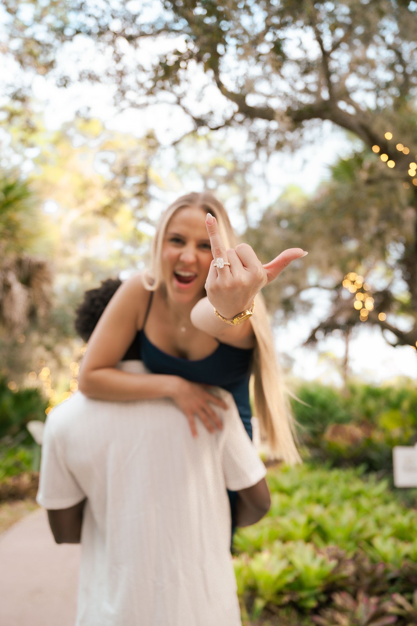 Excited woman showing engagement ring while being carried piggyback by partner in a lush garden setting.
