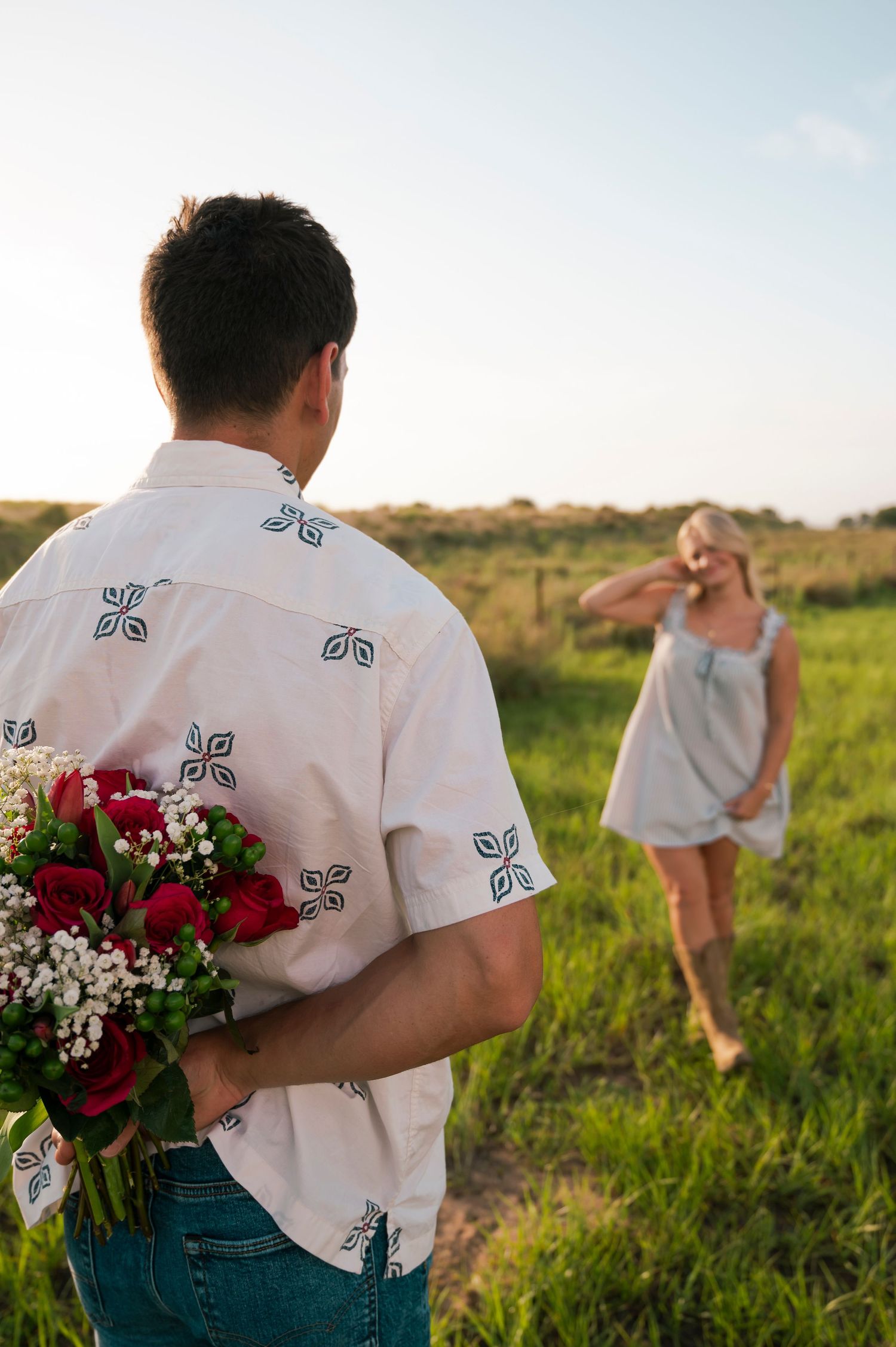 Man hiding red rose bouquet behind his back to surprise a blonde woman in a sunny green field.