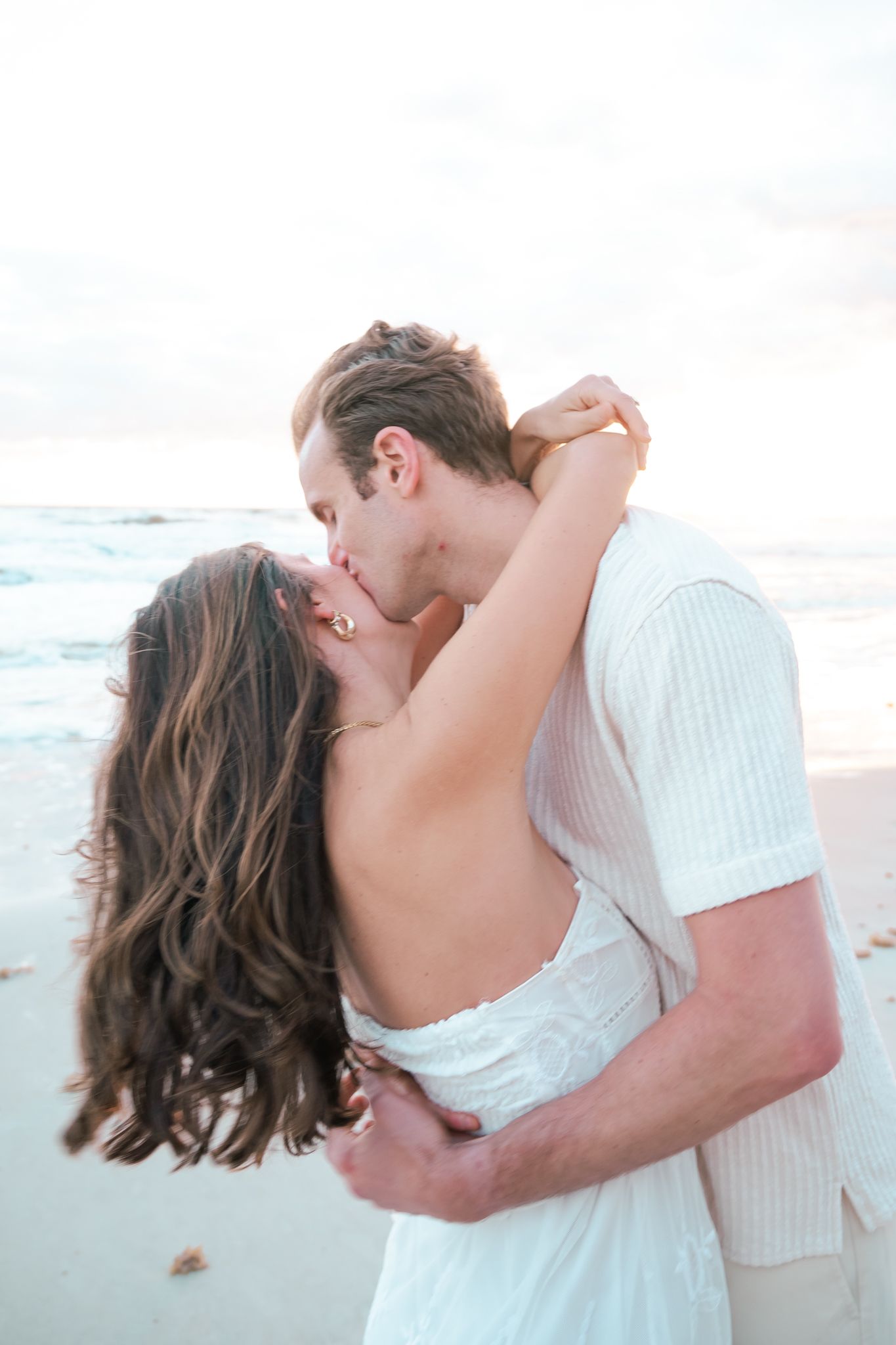 Couple sharing a romantic kiss on a sandy beach, dressed in white, with ocean waves in the background.