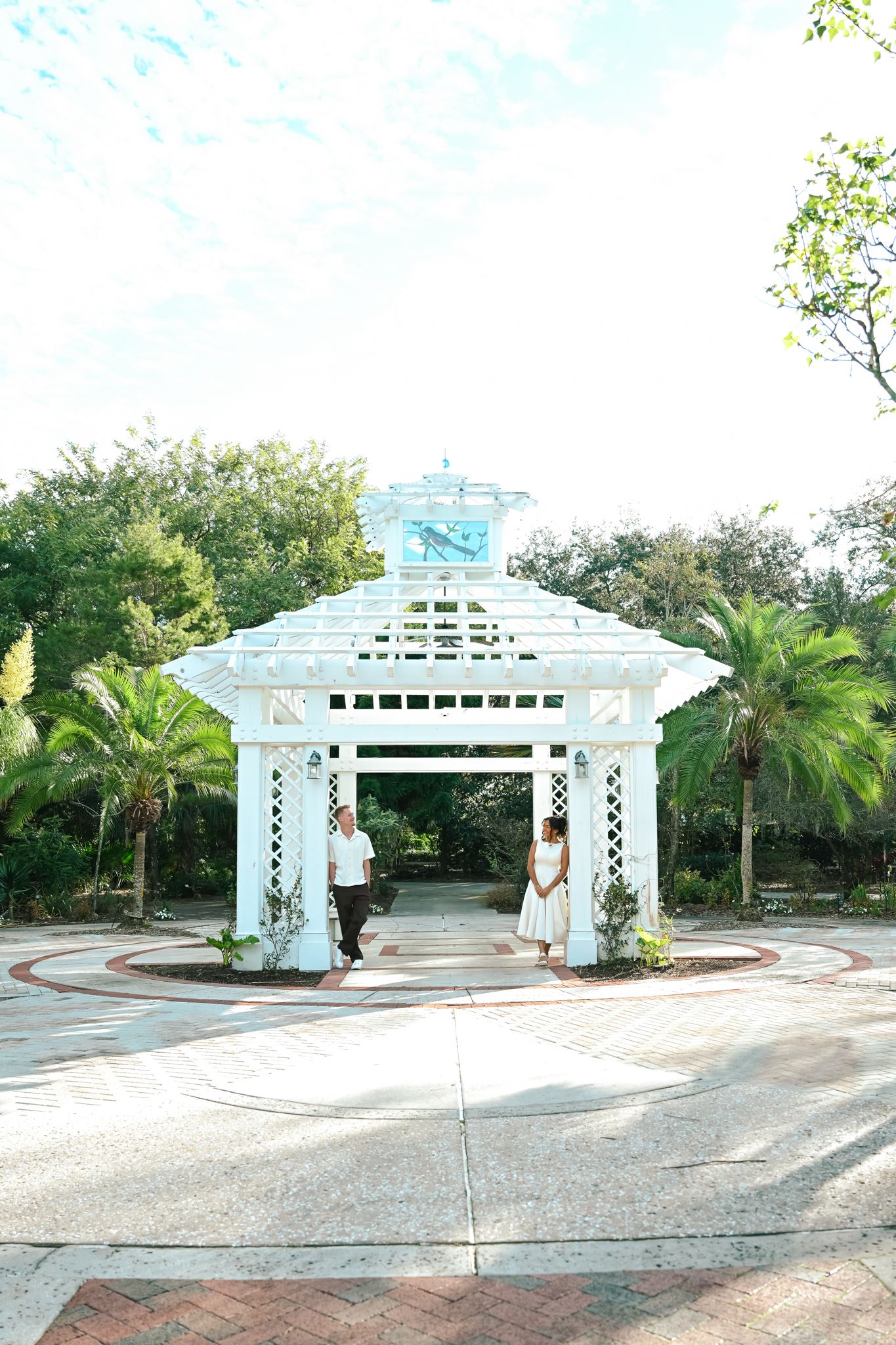 Couple stands under white gazebo surrounded by palm trees and lush greenery at an outdoor garden venue.