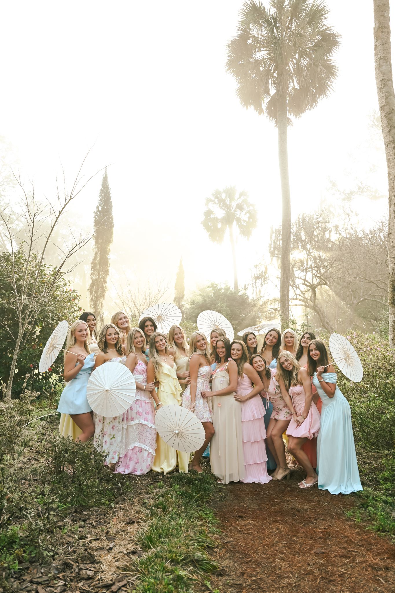 Large bridal party in pastel dresses holding white parasols outdoors with palm trees and golden sunlight.