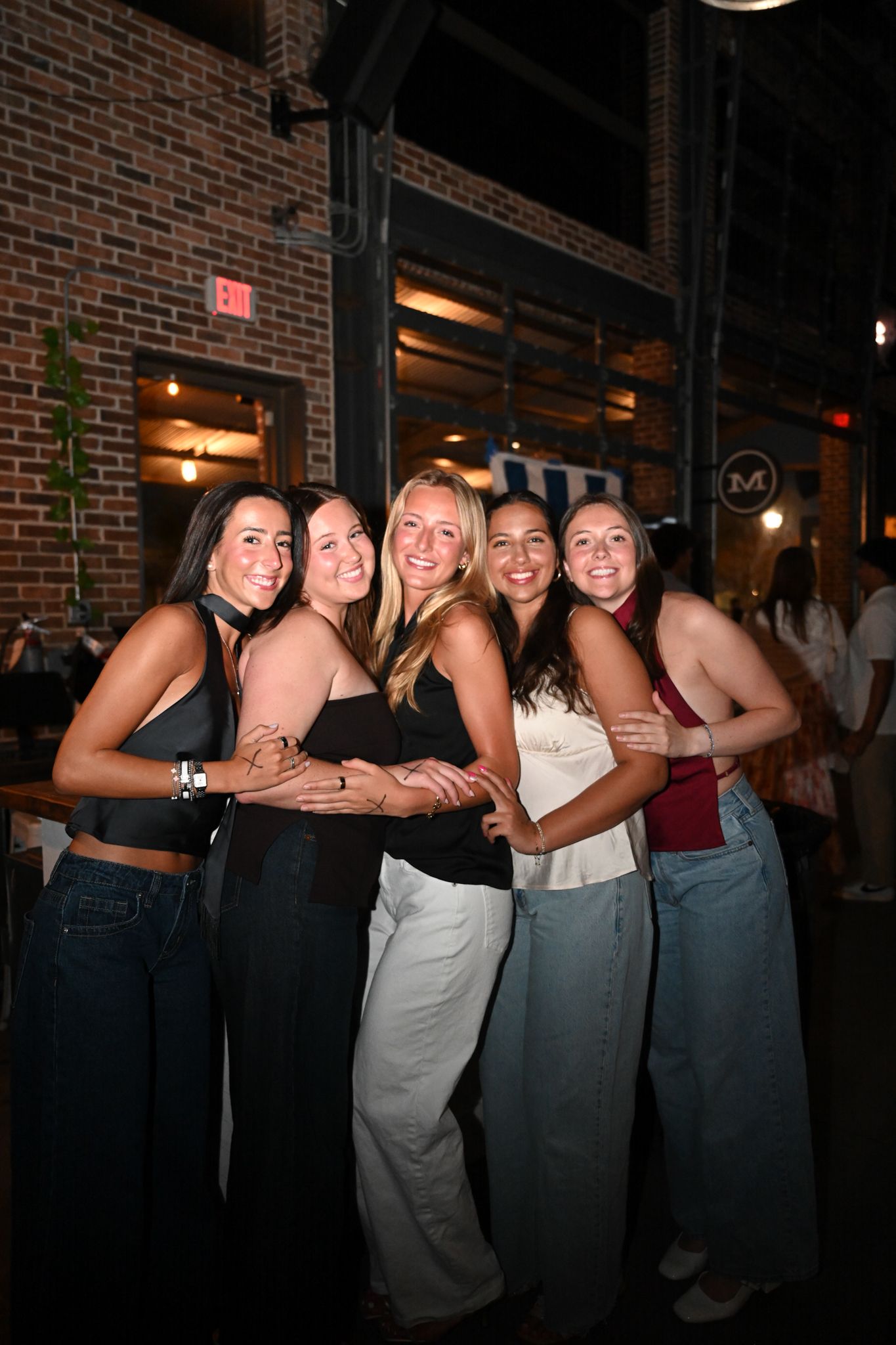 Five young women smiling and posing together outside a brick restaurant at night.