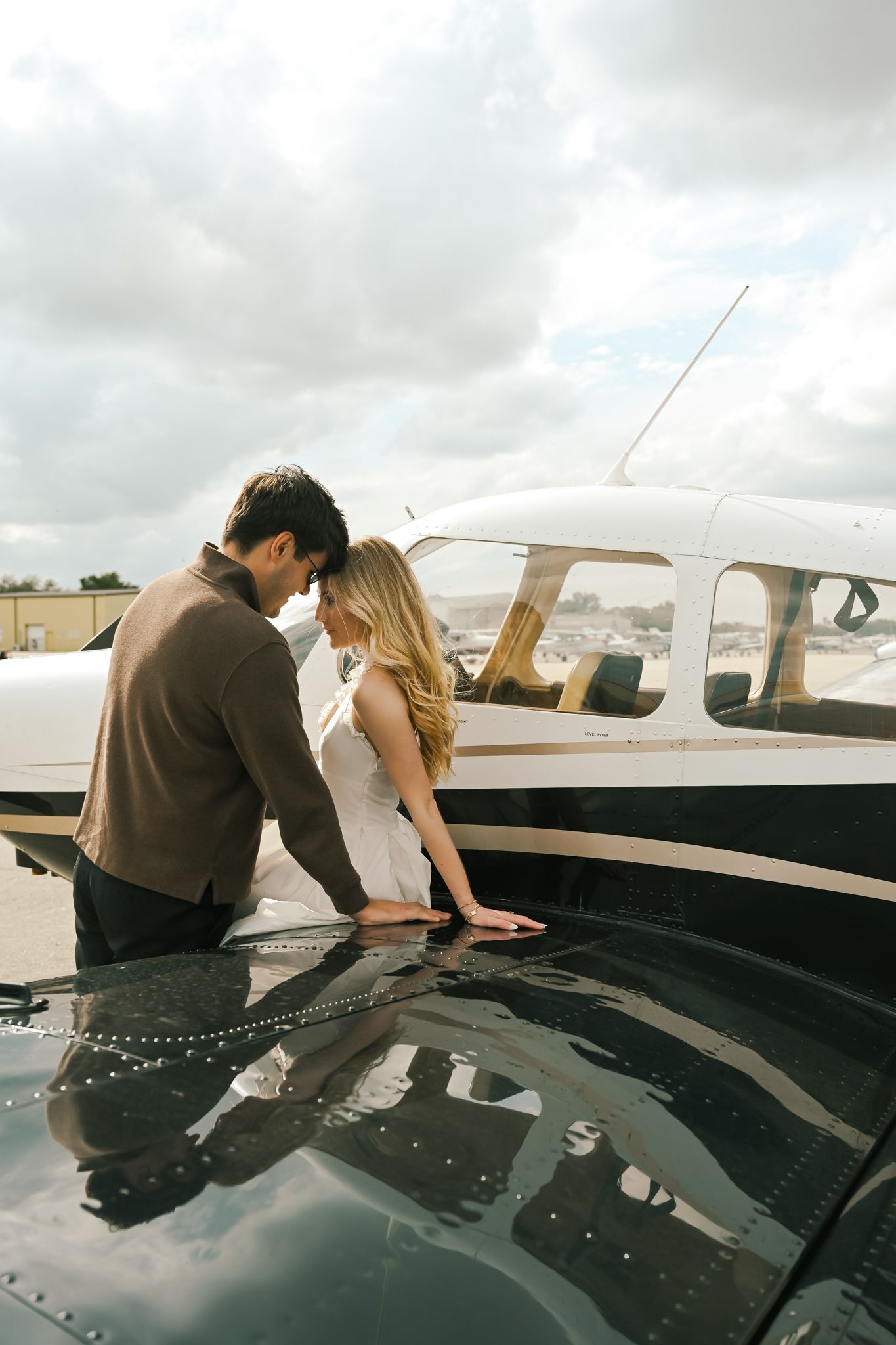 A couple shares a romantic moment leaning close together beside a small private airplane on a cloudy day.