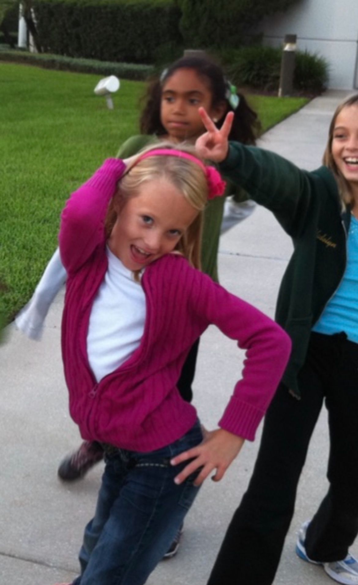 Three girls posing outdoors on a sidewalk, with the front girl in a pink cardigan making a fun pose.