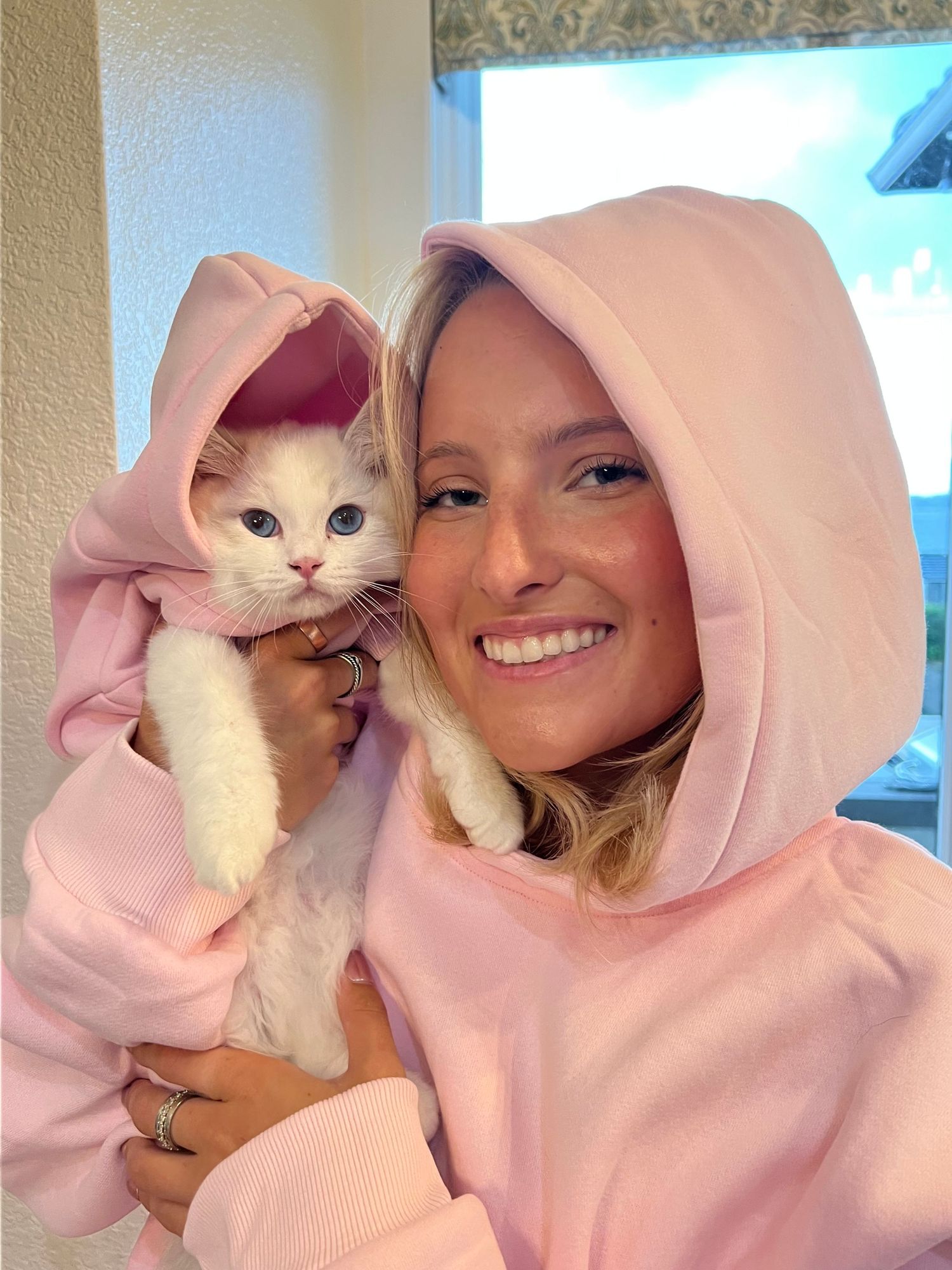 Woman in pink hoodie smiling while holding a white kitten also wearing a matching pink hoodie.