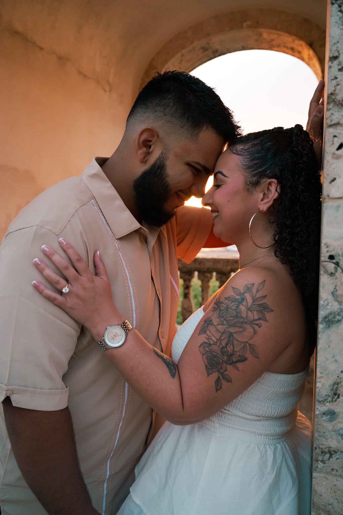 Couple sharing a romantic moment touching foreheads at golden hour, woman with floral shoulder tattoo in white dress.