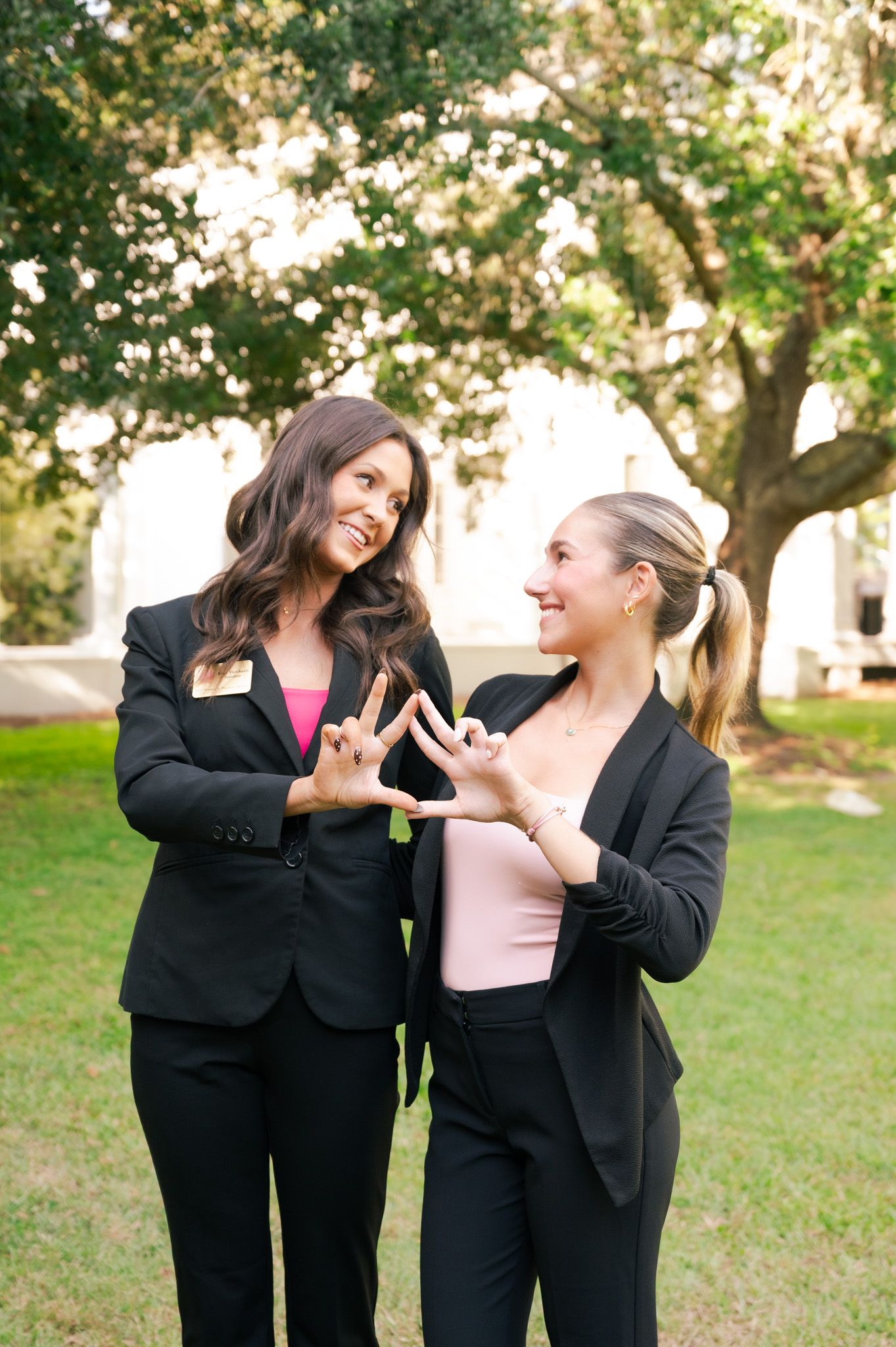 Two women in black blazers smiling and making hand gestures outdoors near trees.