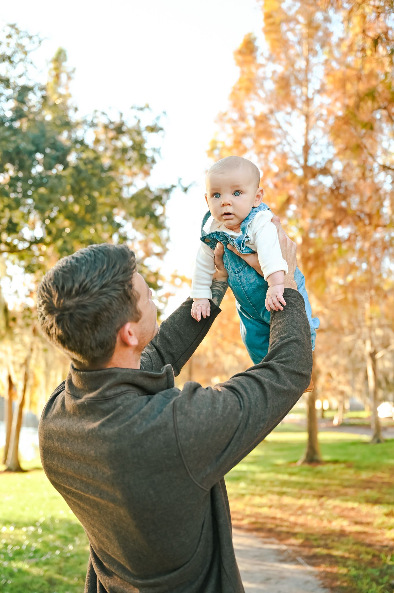 A person lifts a baby high in the air against a backdrop of fall trees.