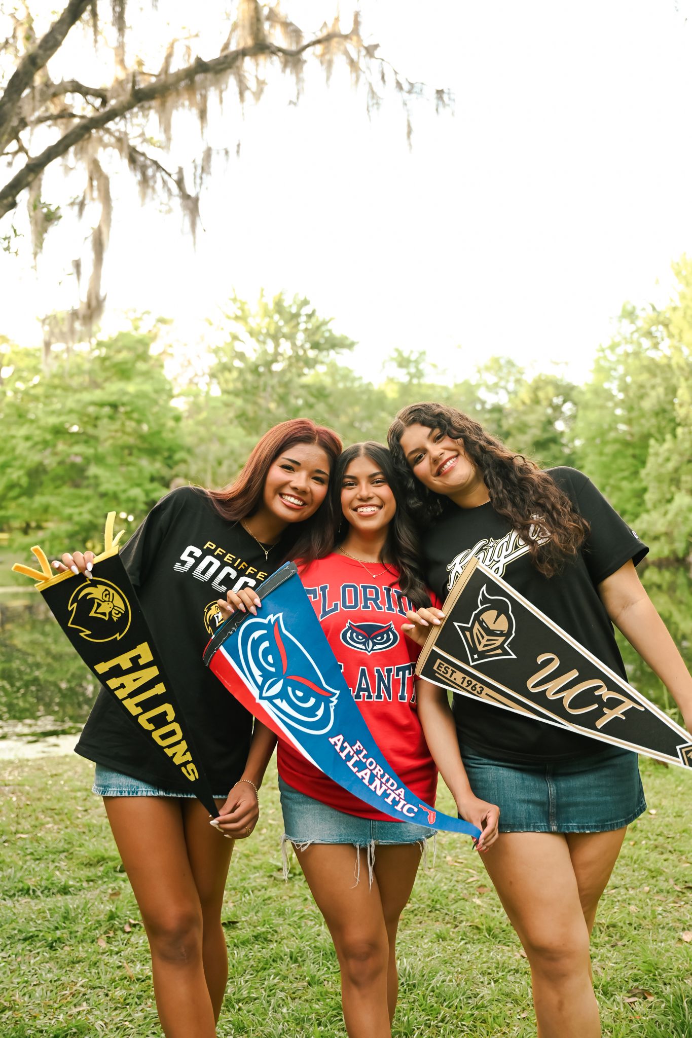 Three young women smile holding college pennants for Falcons, Florida Atlantic, and UCF outdoors.