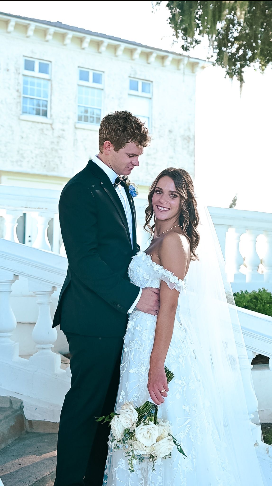 A bride and groom pose together on their wedding day in front of a white colonial-style building.