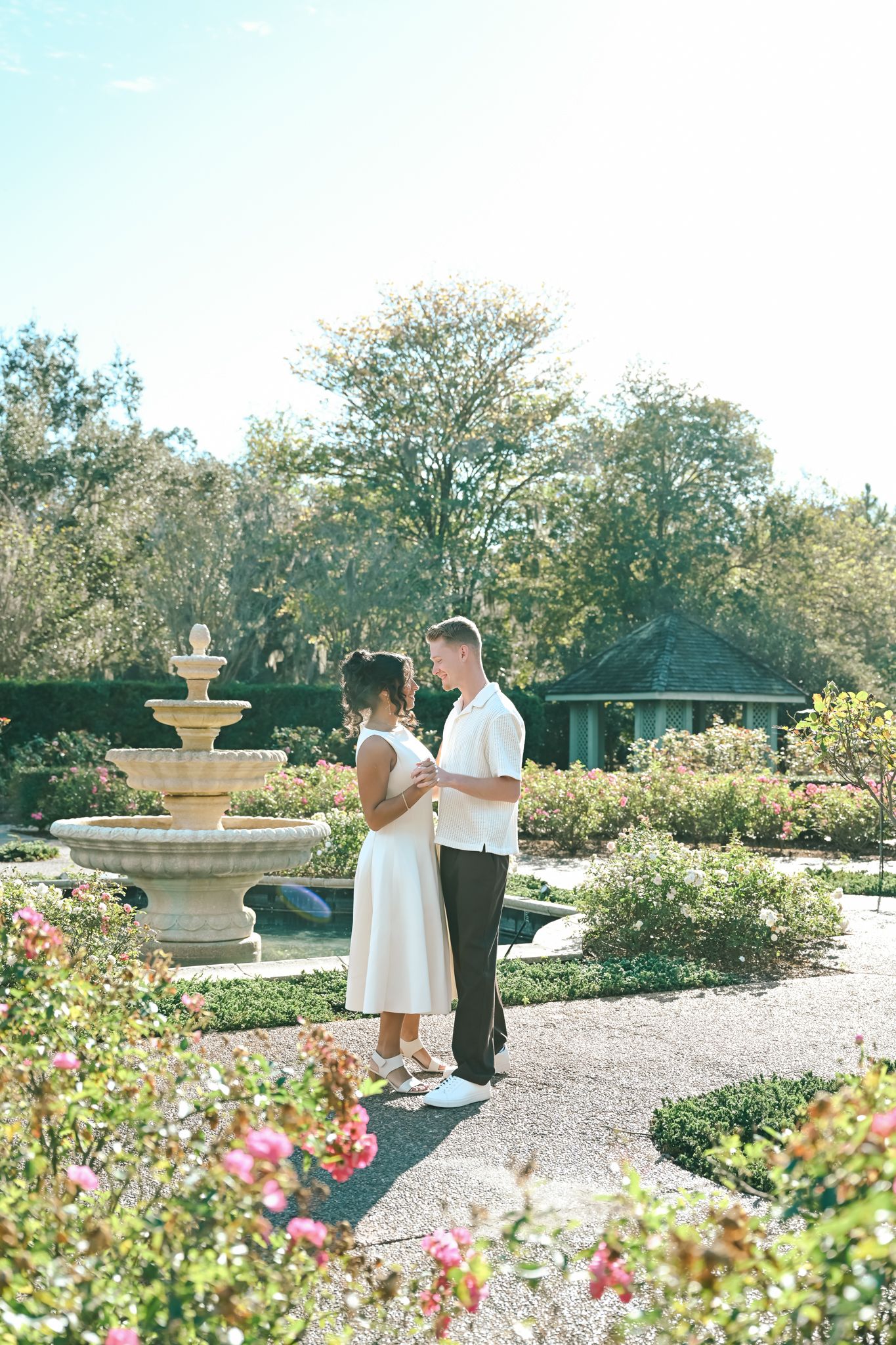 Couple holding hands in a sunlit garden with a stone fountain and blooming roses in the background.