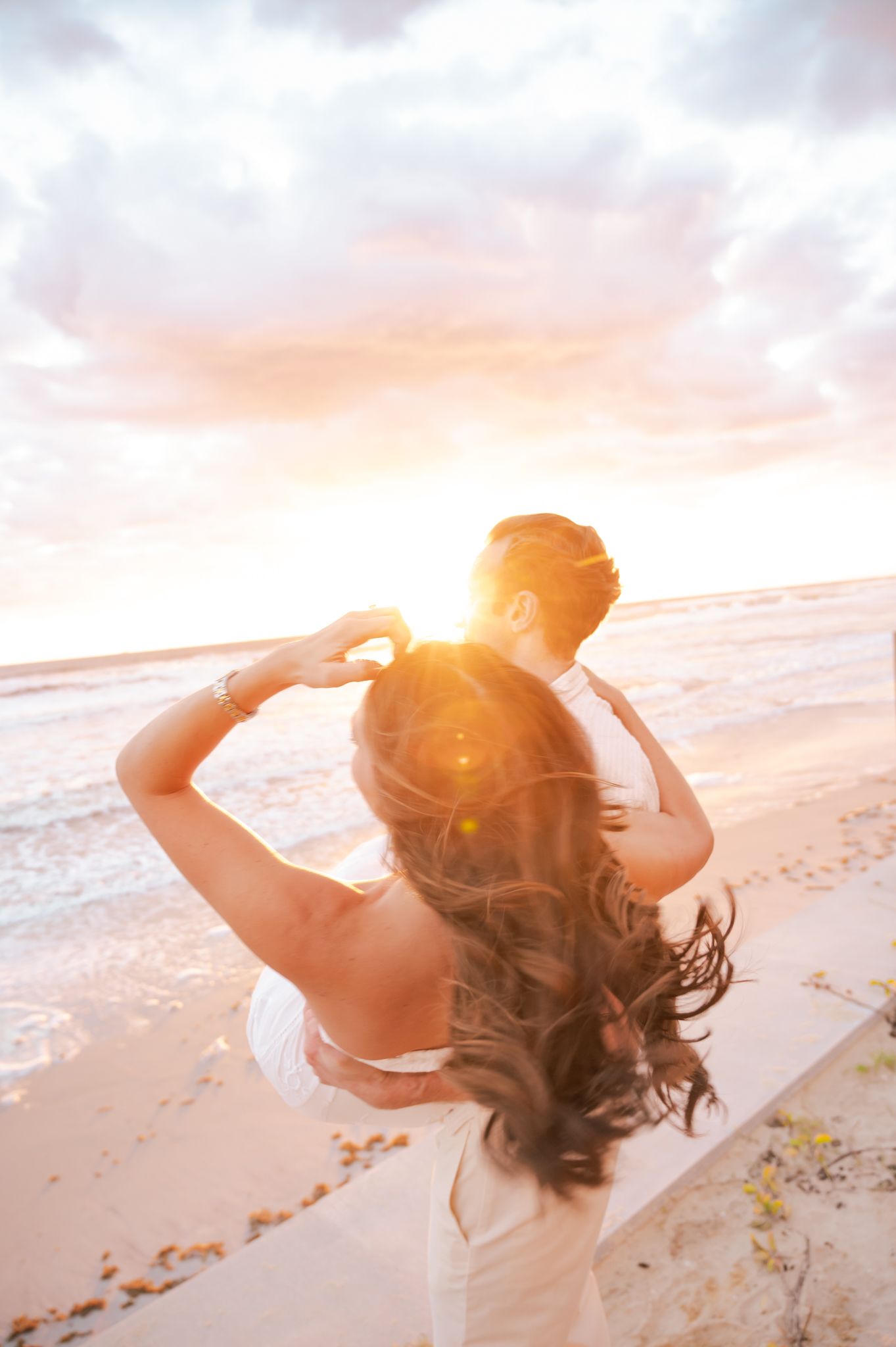 Couple embracing on a sandy beach at golden sunset, woman with long wavy hair facing the glowing horizon.