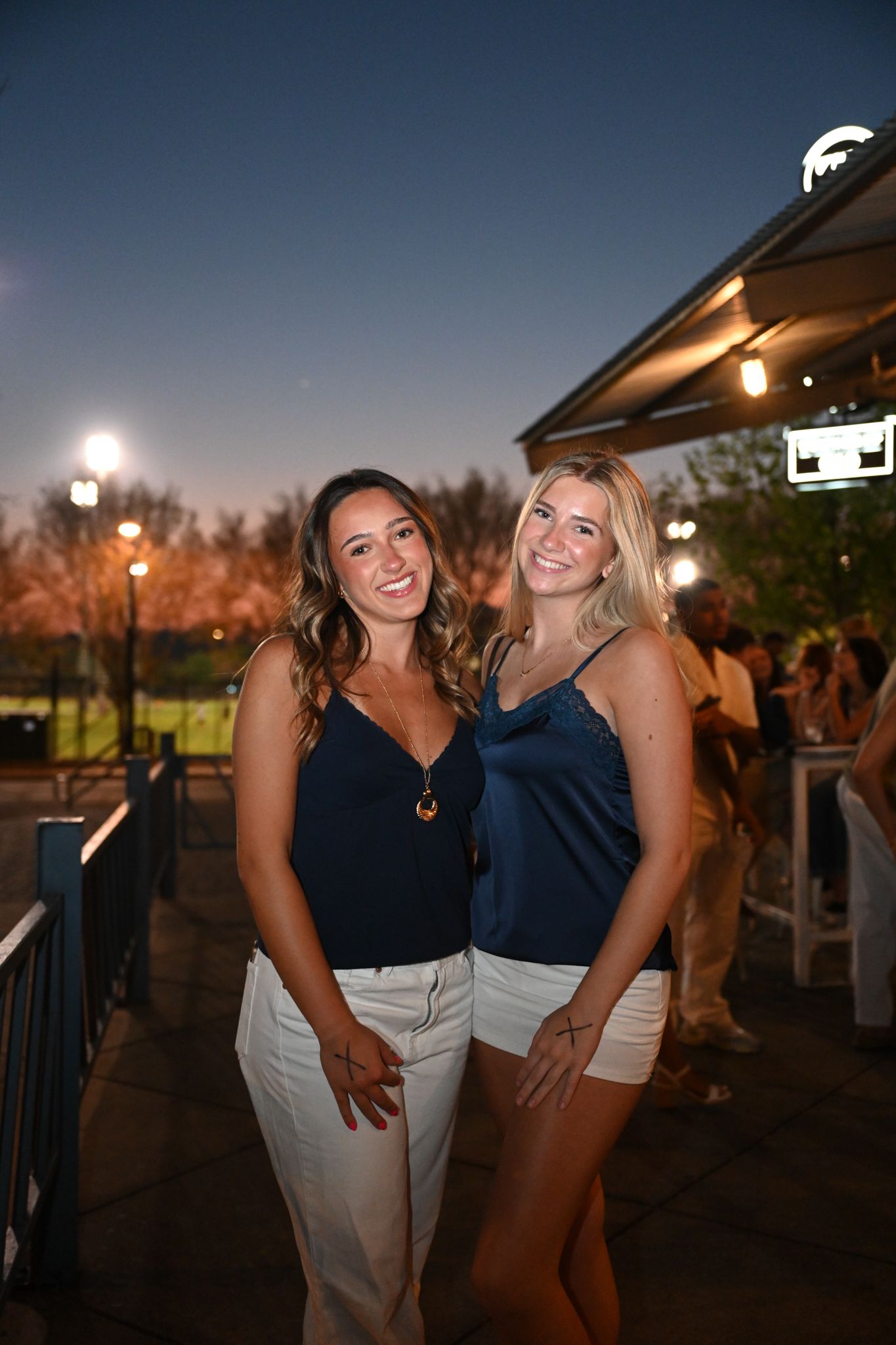 Two smiling women in navy blue tops and white shorts pose together outdoors at a evening social event.