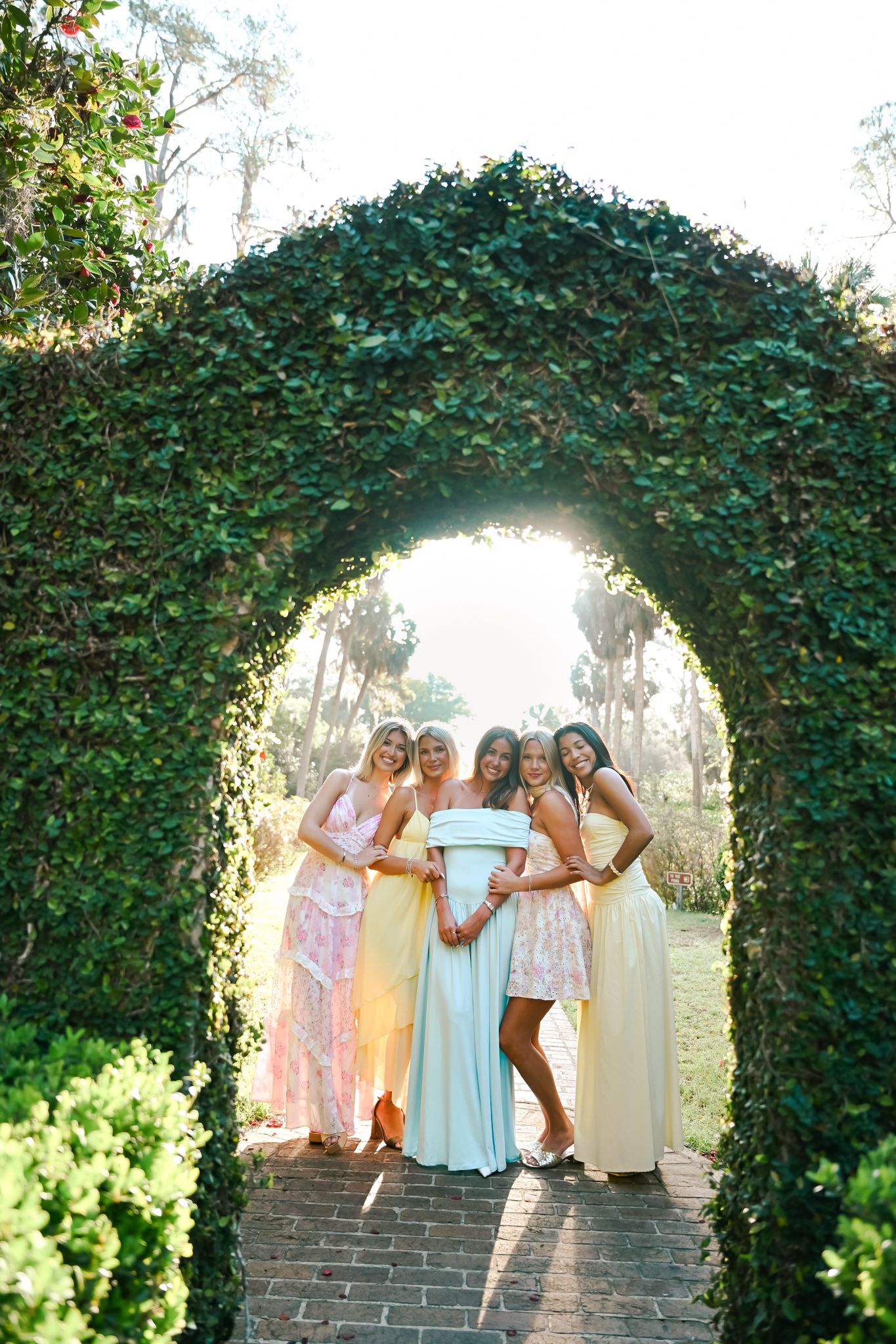 Bridesmaids in pastel gowns pose with bride under ivy-covered garden arch on brick pathway.