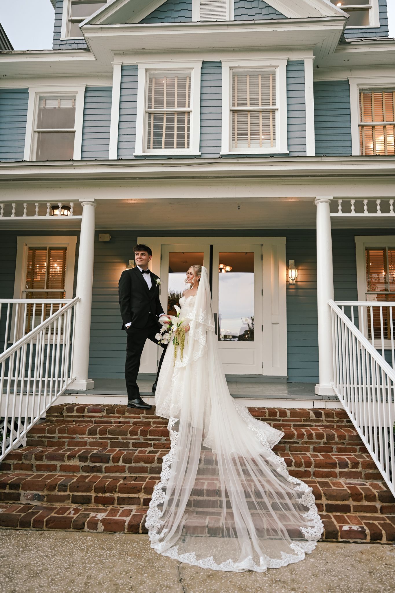 Bride and groom pose on brick steps of a blue Victorian house, her lace cathedral veil cascading down the stairs.