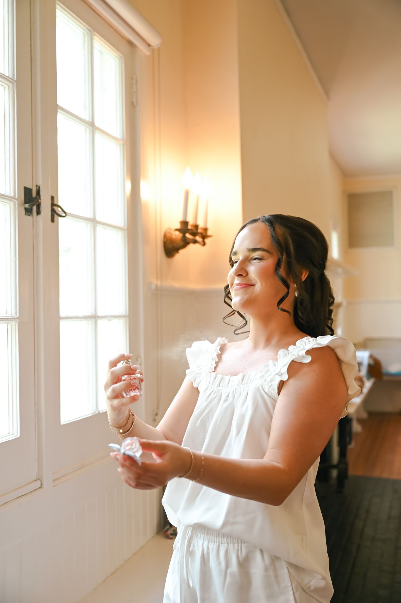 Smiling bride in white dress holding perfume bottle near window in elegant room.