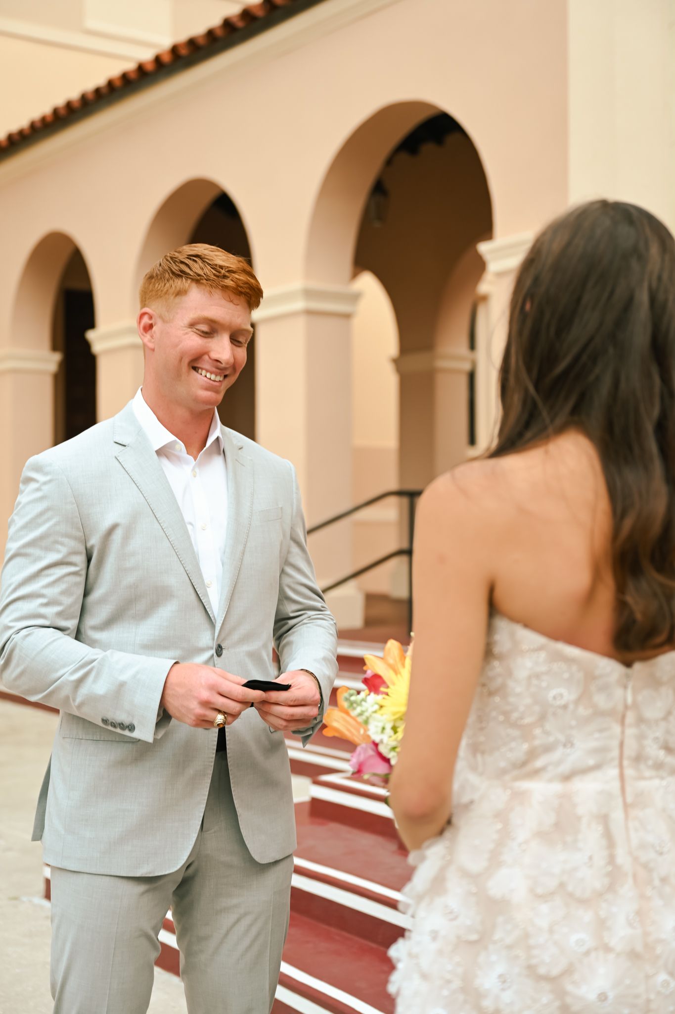 Smiling redhead groom in gray suit reads note while bride in lace dress stands before him at elegant venue.