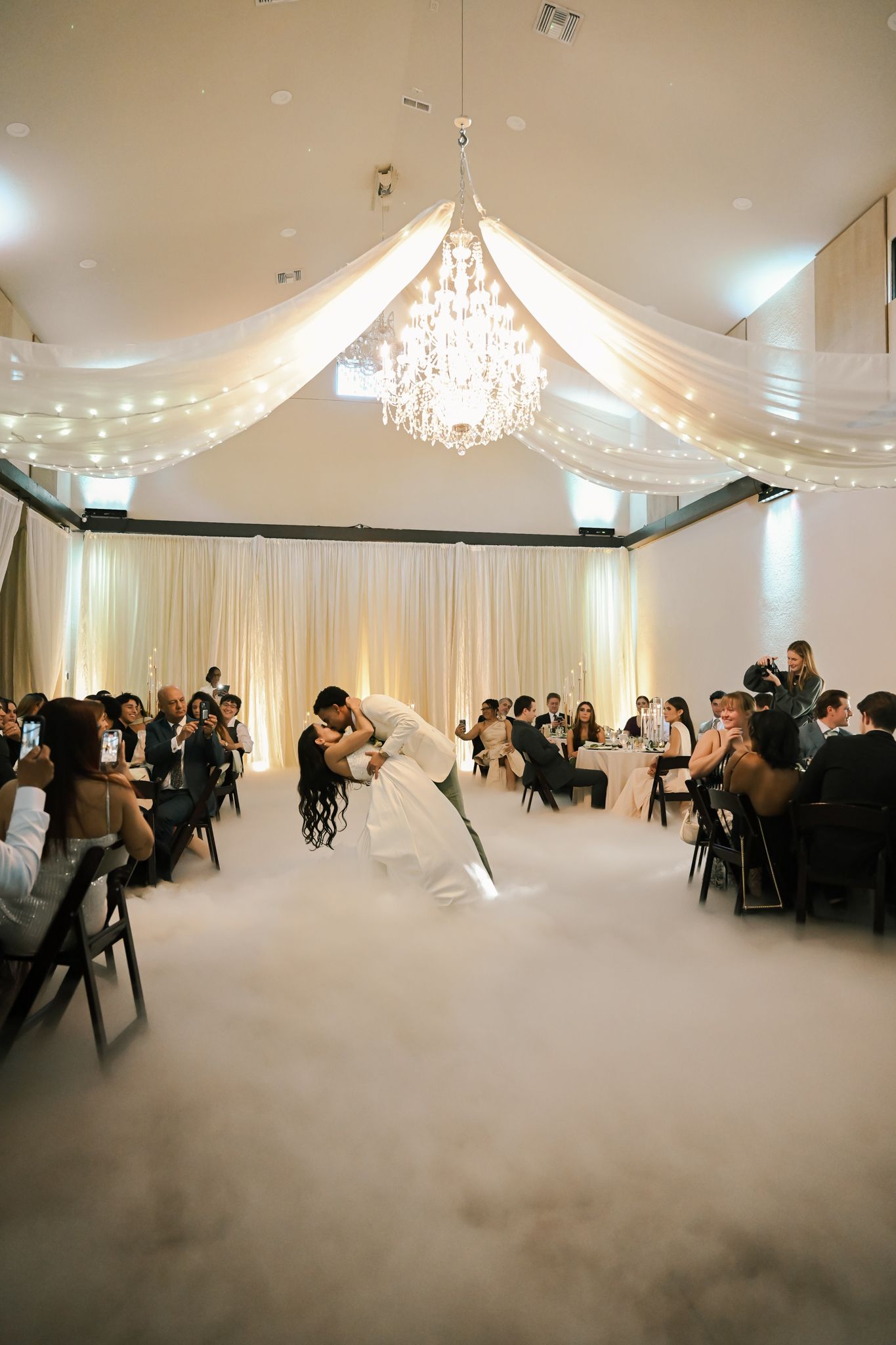 Bride and groom share a romantic dip kiss on a foggy dance floor beneath a chandelier at an elegant wedding reception.