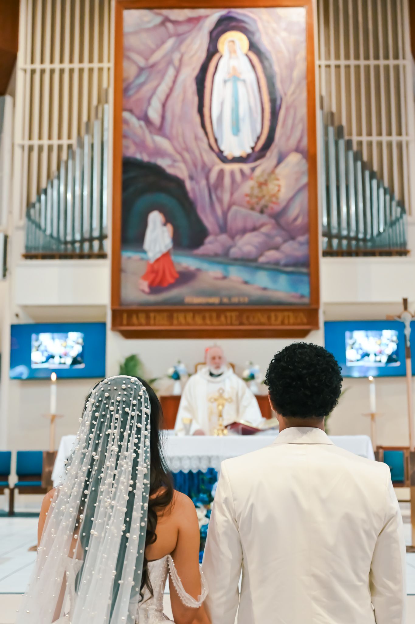 Bride and groom stand before a priest at a Catholic wedding altar beneath a painting of the Immaculate Conception.