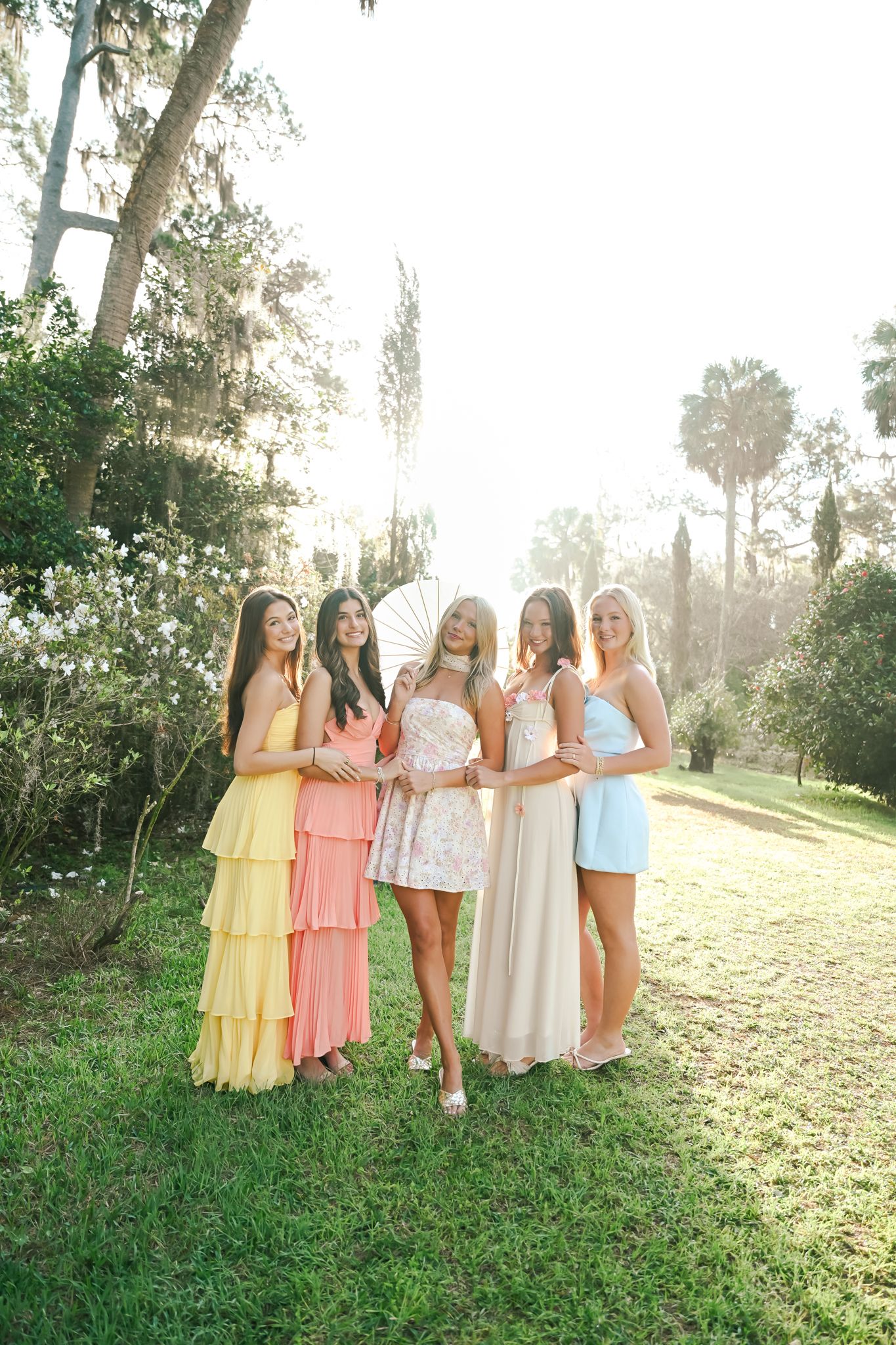Five women in pastel dresses pose together outdoors in a sunlit garden with lush green trees.