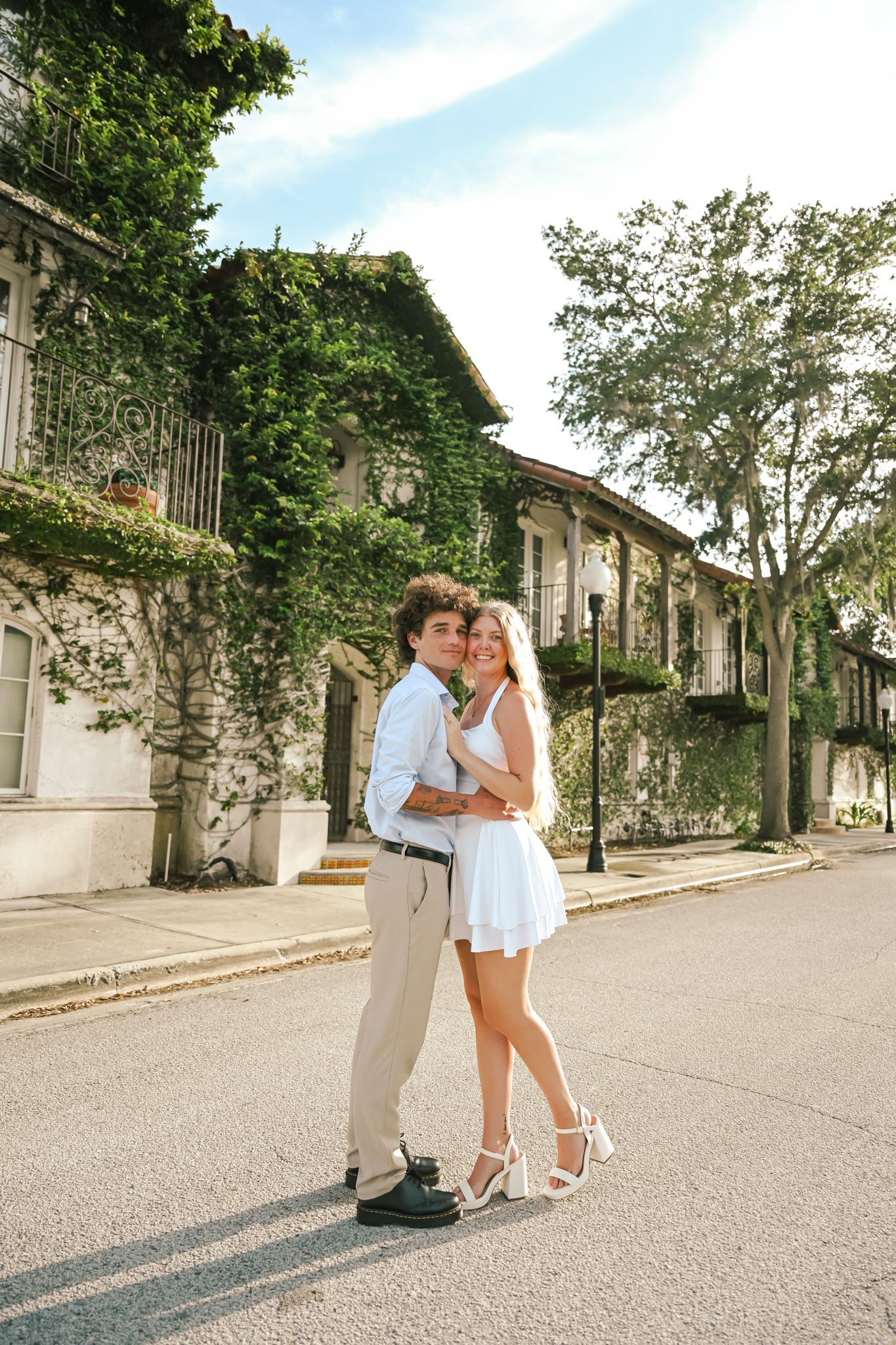A couple embraces on a sunny street lined with ivy-covered buildings and trees in a charming neighborhood.