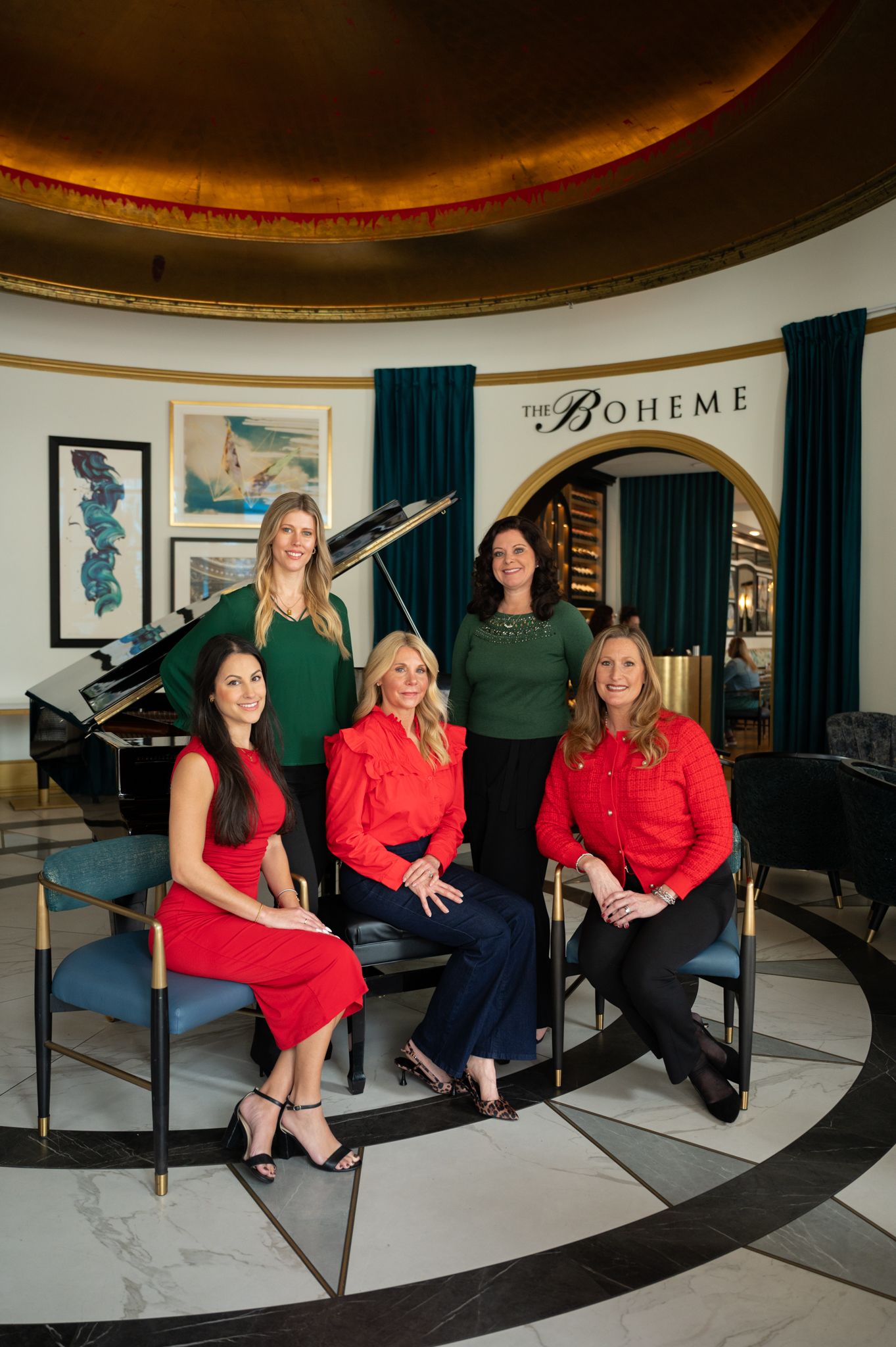 Five professional women posing elegantly at The Boheme venue, featuring a grand piano and teal drapes.