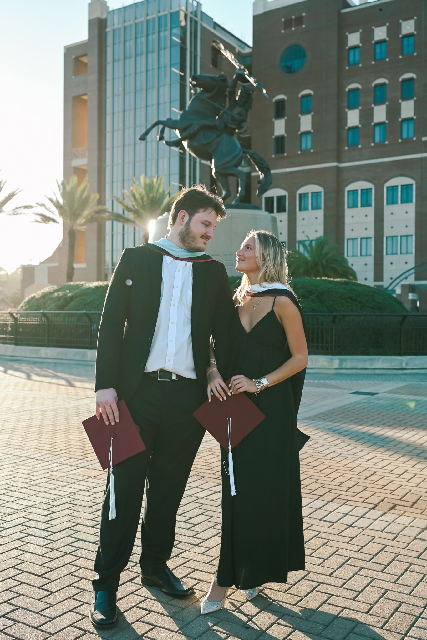 Two graduates in black gowns holding diplomas smile at each other in front of a university statue on a sunny day.