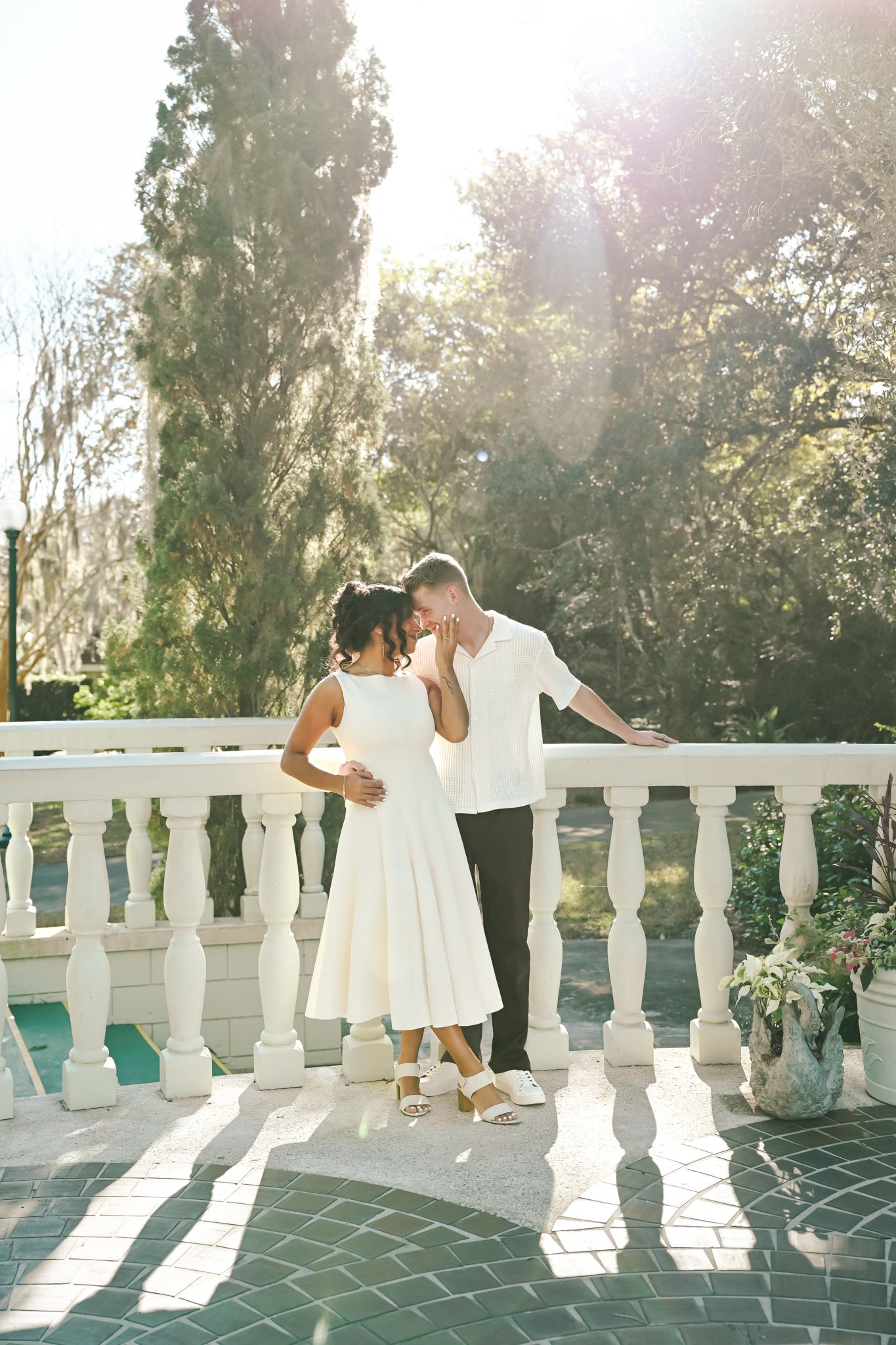 Couple in white outfits sharing a romantic moment on a sunlit balcony surrounded by lush green trees.