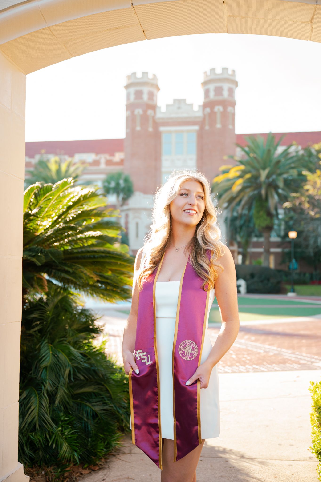 Graduate in burgundy stole poses near palm trees and historic brick buildings.