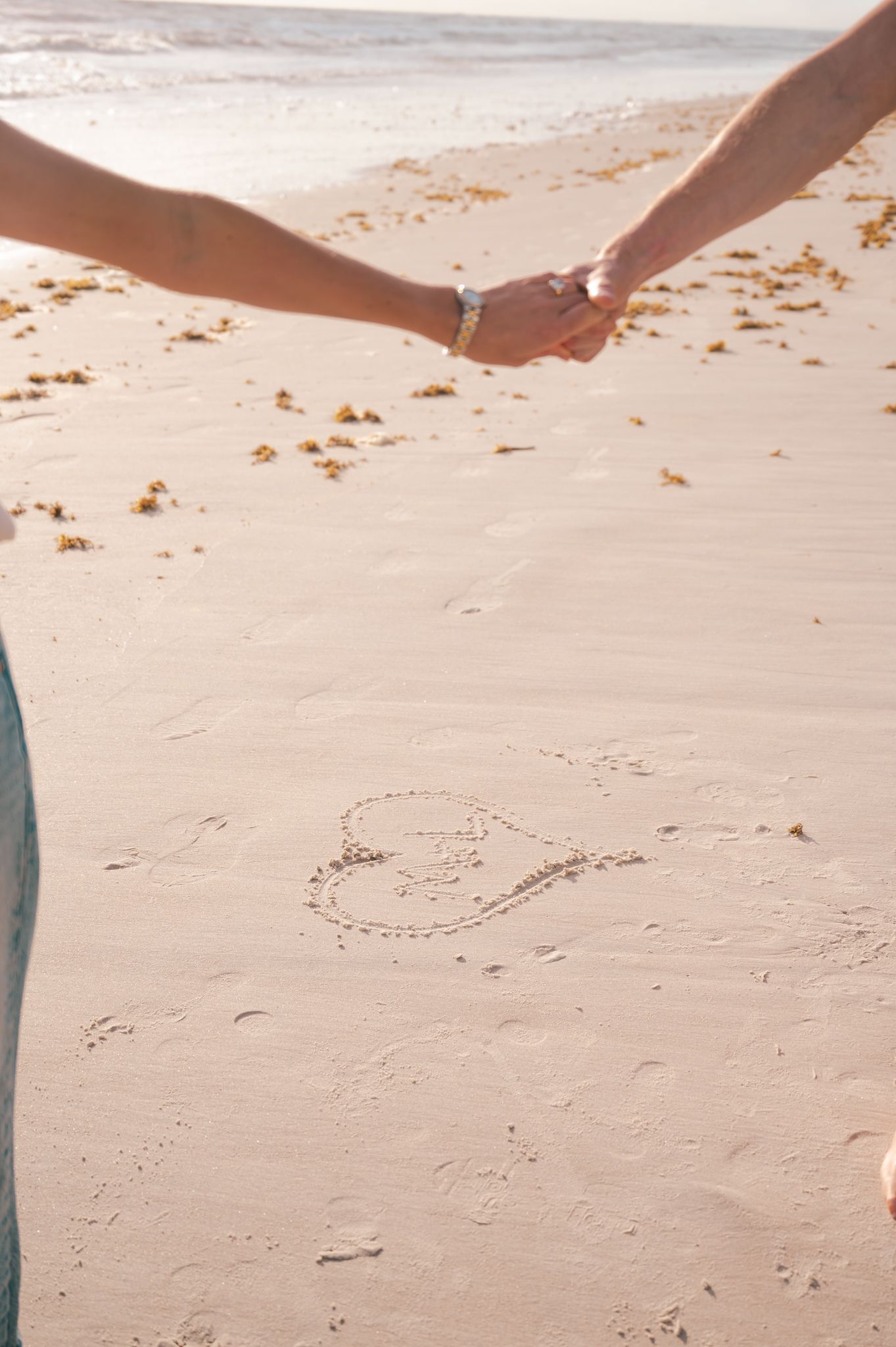Couple holding hands on sandy beach with a heart drawn in the sand near the ocean shoreline.