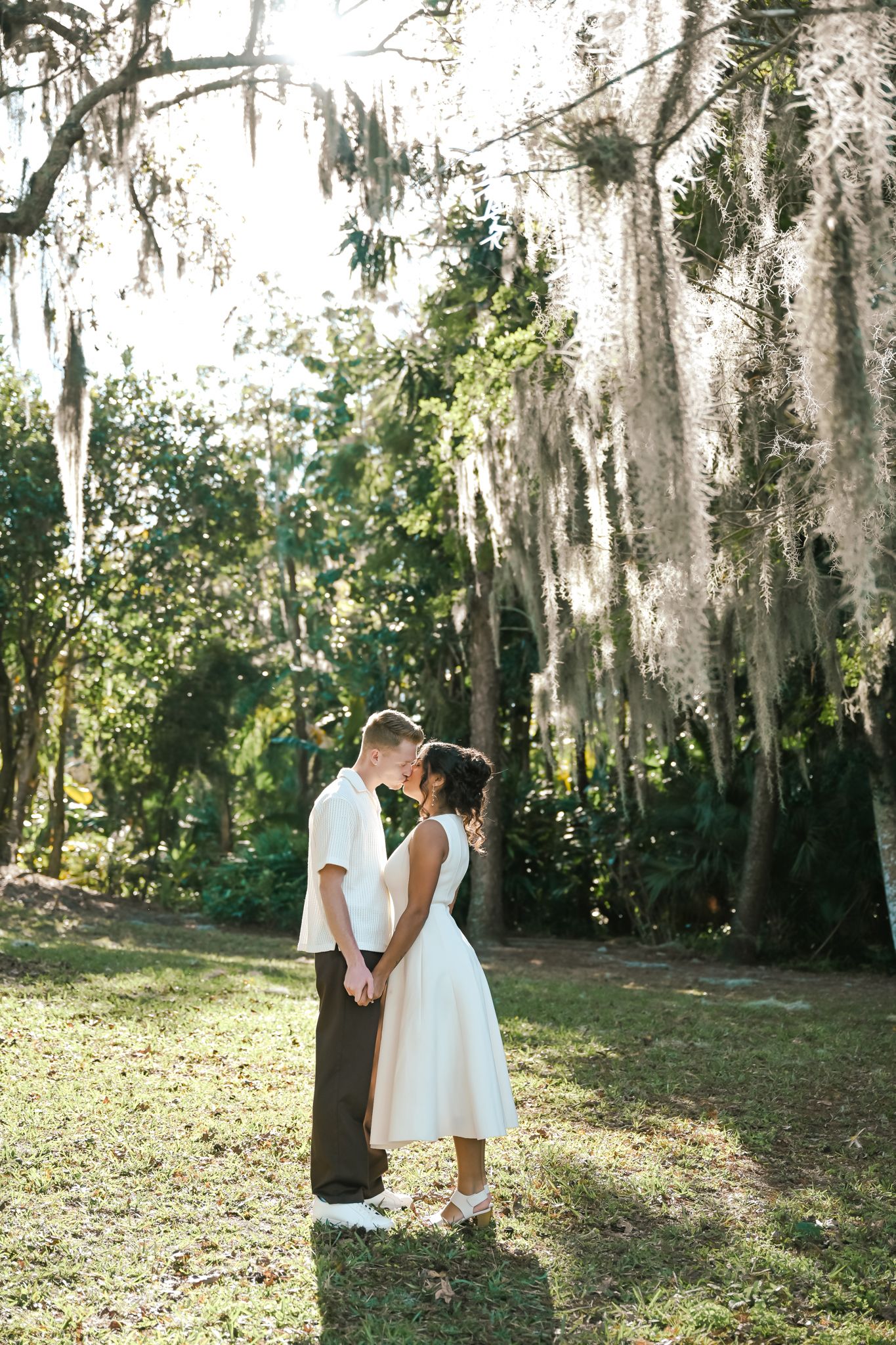 Couple sharing a romantic kiss under Spanish moss-draped trees in a sunlit garden, bride in white midi dress.