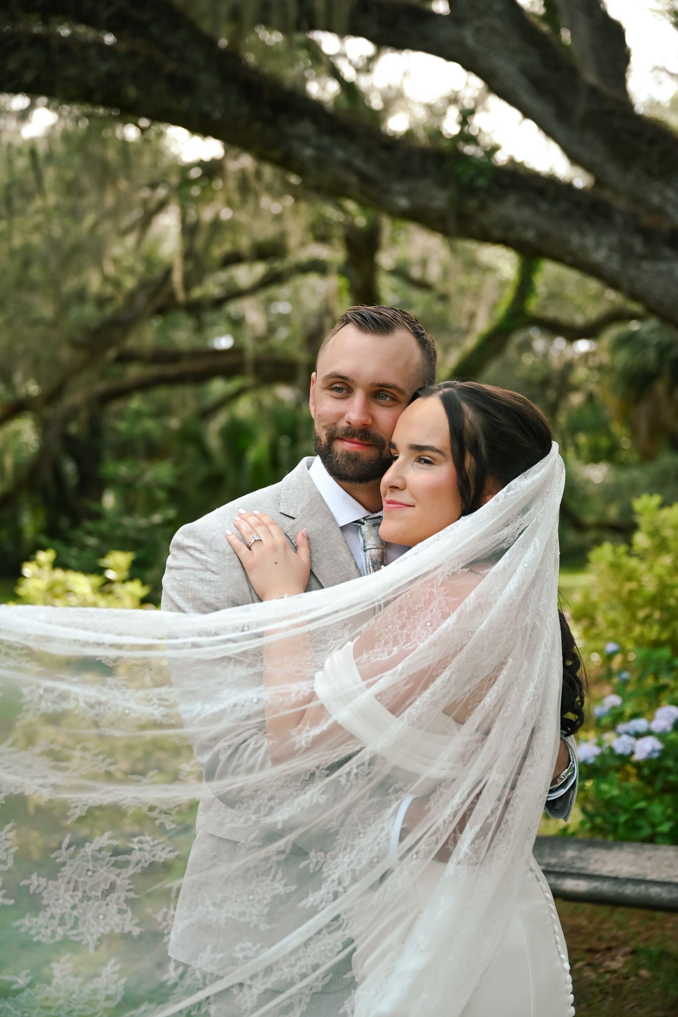 Bride and groom embrace outdoors under moss-covered oak trees, her lace veil flowing around them in a romantic garden wedding portrait.