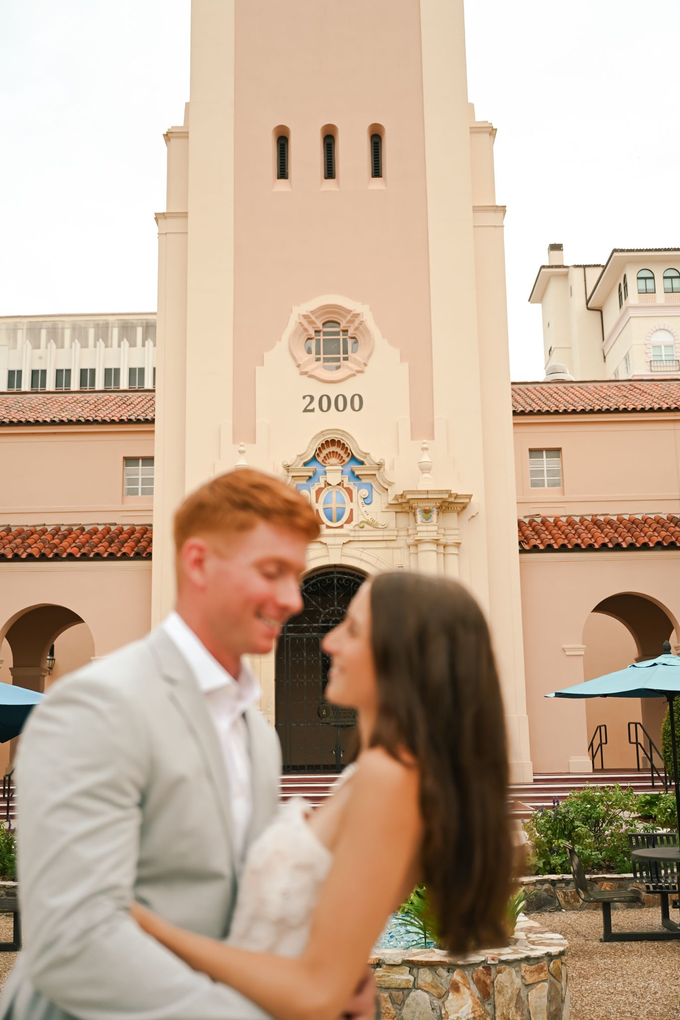 Couple embracing in front of a pink Spanish-style building with a tower marked 2000, dressed in wedding attire.