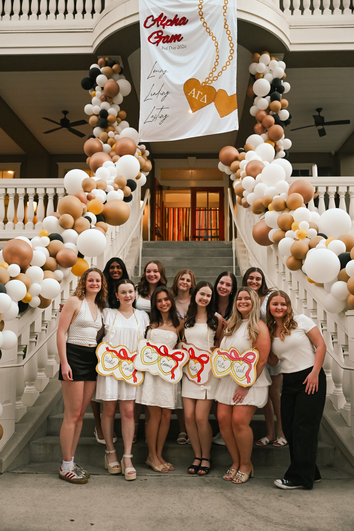 Group of young women in white dresses pose on decorated stairs with balloons at Alpha Gamma Delta sorority event.